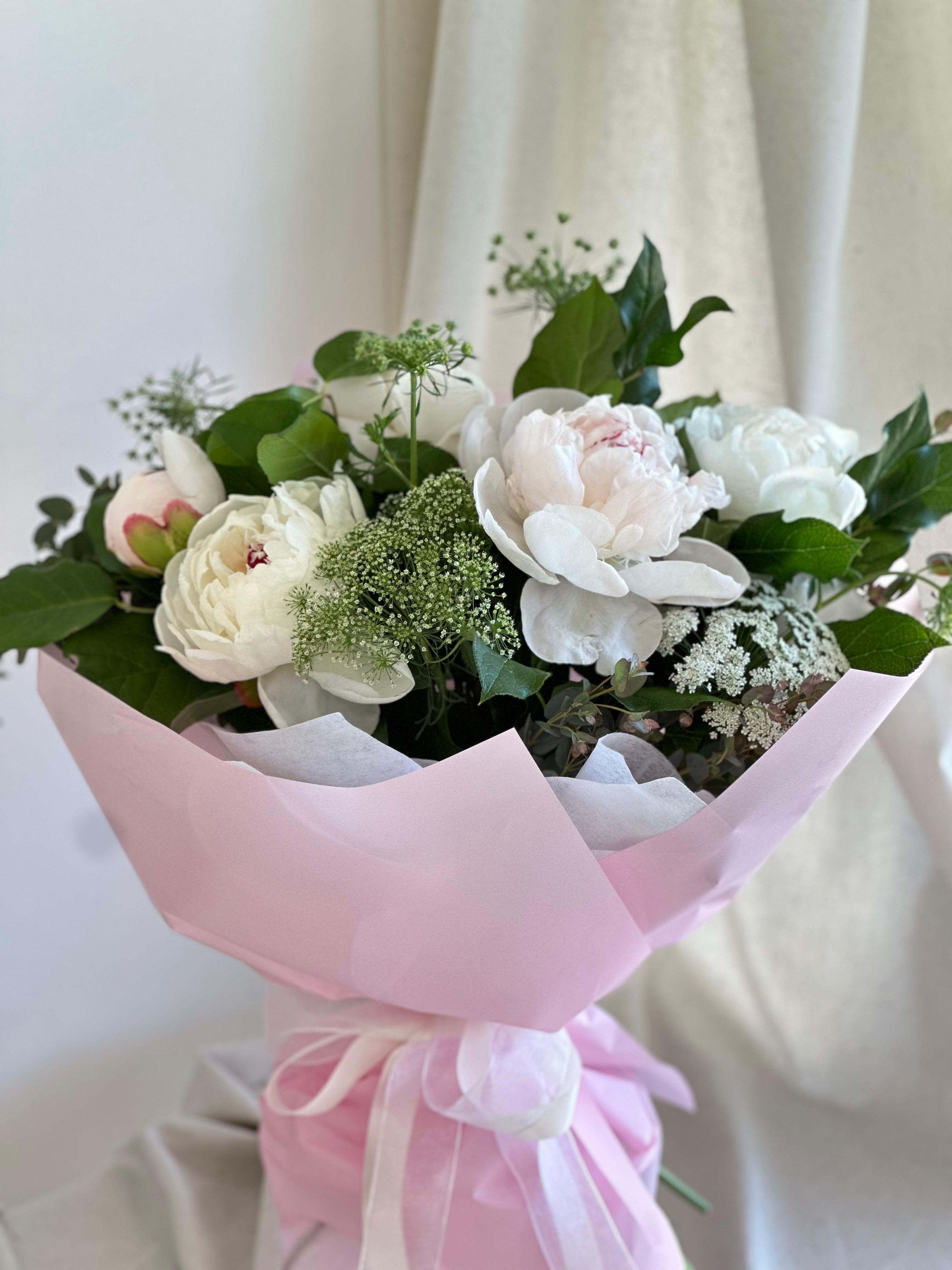 Peonias in Blanco  - A round bouquet of lt. pink peonies with plenty of greenery and fillers, wrapped in pink paper.   Flower types may vary depending on the season.