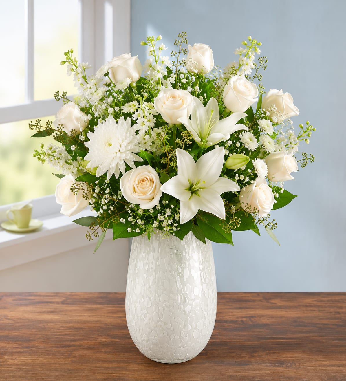 Royal Harmony - Pure white lilies and roses arranged with crisp greenery create a serene, comforting presence-perfect as a sympathy arrangement or a thoughtful New Baby gift that conveys calm, hope, and gentle care. This upright floral arrangement also makes an elegant choice for funerals, memorial services, or Hanukkah centerpieces, and its quiet palette translates well for winter holidays like Christmas without feeling seasonal-only.  Creamy white roses, fragrant white lilies, and white chrysanthemums are gathered with airy white filler blooms and bright green foliage in a white ceramic vase for a clean, rounded silhouette. The design reads formal yet soft: full and layered with delicate branching accents that add lightness and texture. Available sizes are generous and upright; lilies may arrive in bud form and will open over the following days to extend the display.  This arrangement is the kind of gift that puts the recipient at ease-whether you're honoring a life, welcoming a new baby, or offering comfort from afar. Each piece is hand-designed by our florists and vase style may vary slightly by availability; care is simple-fresh water and a cool spot help blooms last longer.