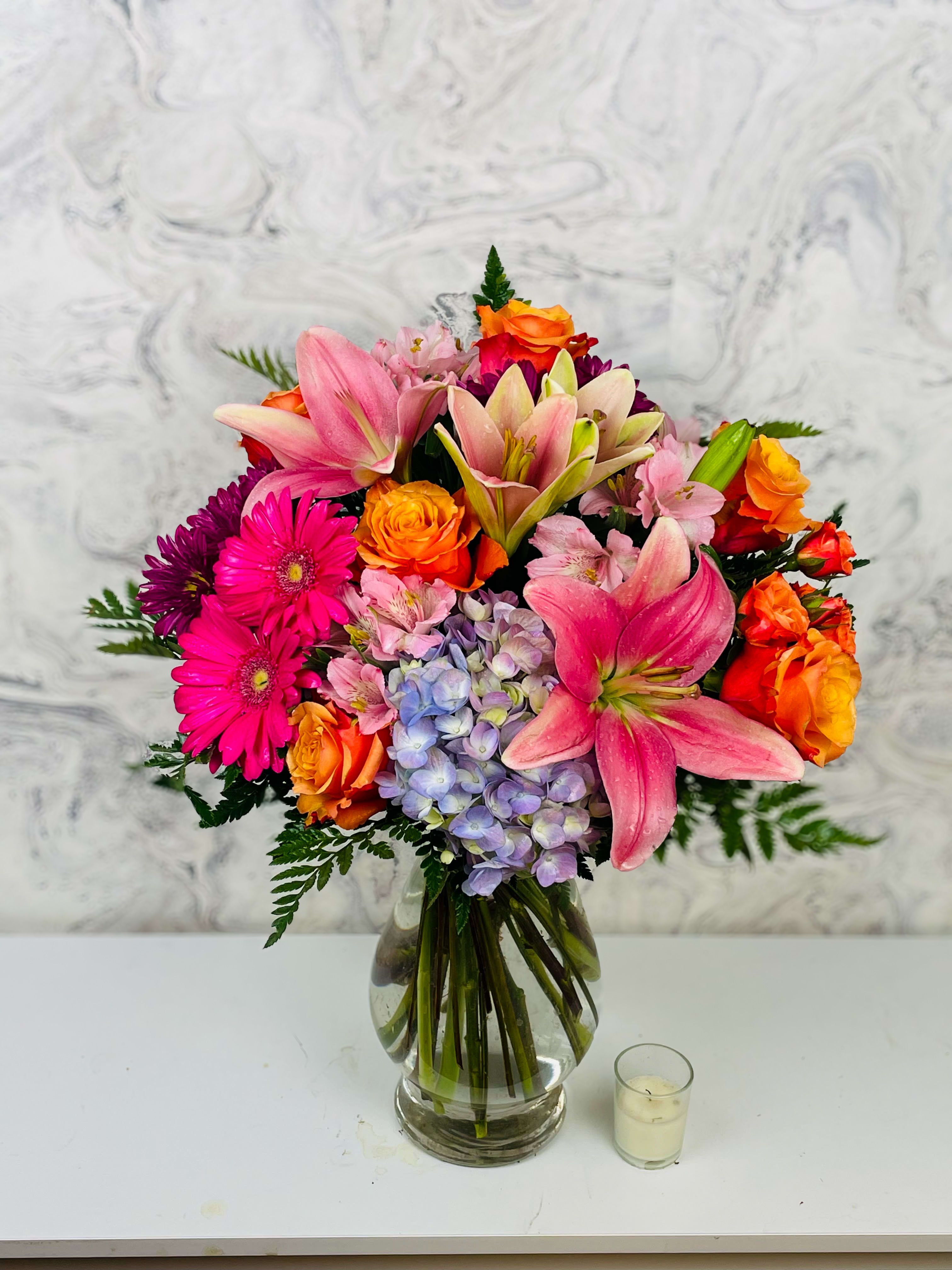 Mixed bouquet of pink lilies, orange roses, and purple flowers in a glass vase