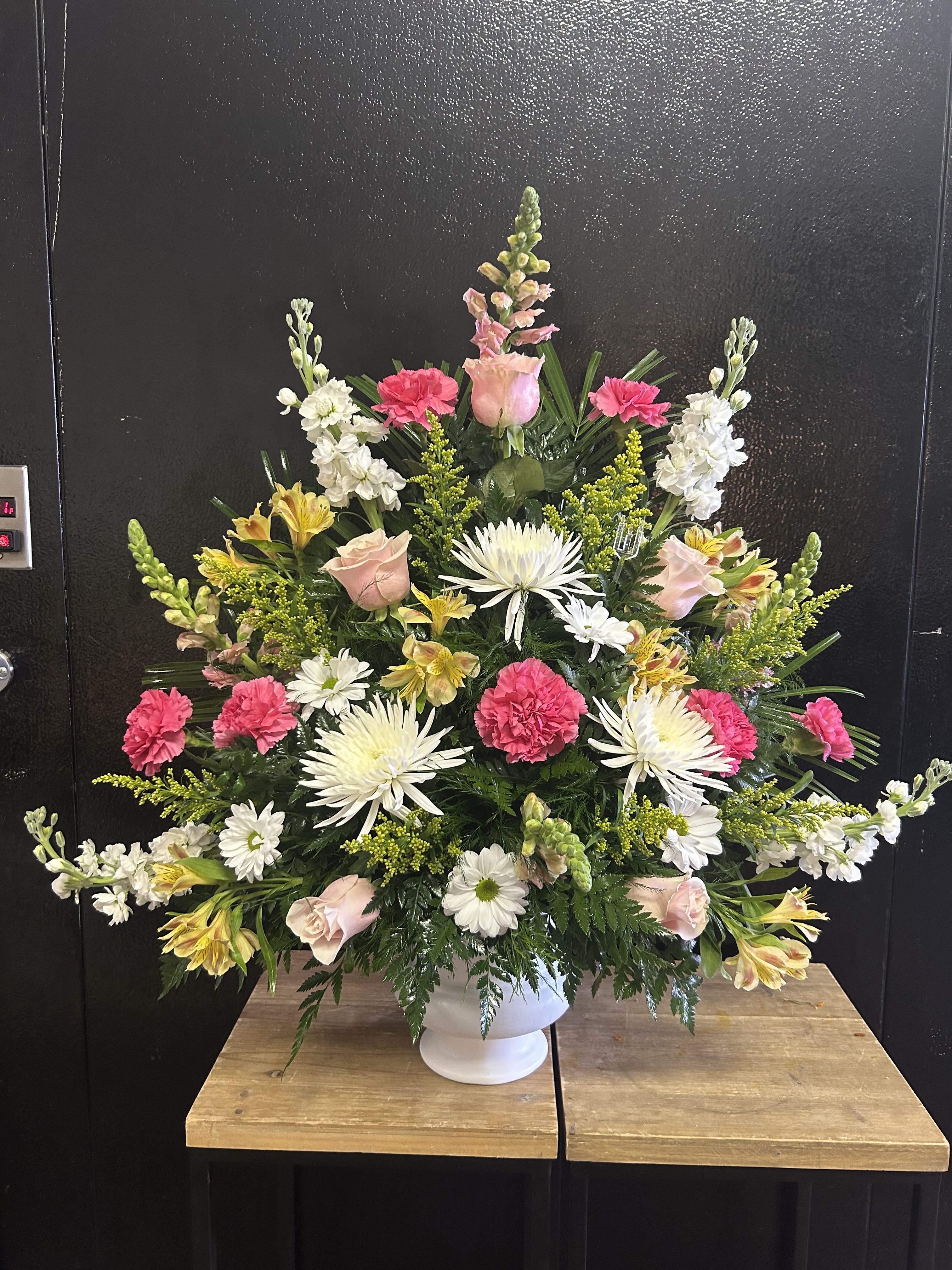 Warm Embrace Bouquet  - A dignified standing sympathy arrangement that comforts with soft color and classic form, perfect as a funeral spray or memorial tribute. The Warm Embrace bouquet pairs traditional white lilies, roses, chrysanthemums, and stock for a serene, formal silhouette, while pale pink roses, yellow alstroemeria, and white daisies add gentle warmth-an arrangement designed to make the person you're honoring feel respectfully remembered.  This design is presented as a tall, tiered display: one composition sits on a sturdy wire easel for a classic standing spray profile, and another fills a white pedestal vase with a fan-like, symmetrical shape. Expect layered greenery for fullness, tall snapdragons for vertical presence, and a dense, balanced distribution of blooms from top to bottom. The palette of pink, yellow, white, and mixed tones reads calm yet comforting, suitable for a funeral service or visitation.  Choose this piece when you want a meaningful, composed statement of sympathy that reads both formal and tender. Sizes are substantial to suit sanctuary and chapel settings; stems are arranged for stability on the easel or in the pedestal vase-no special care is needed beyond keeping blooms in a cool place until the service.