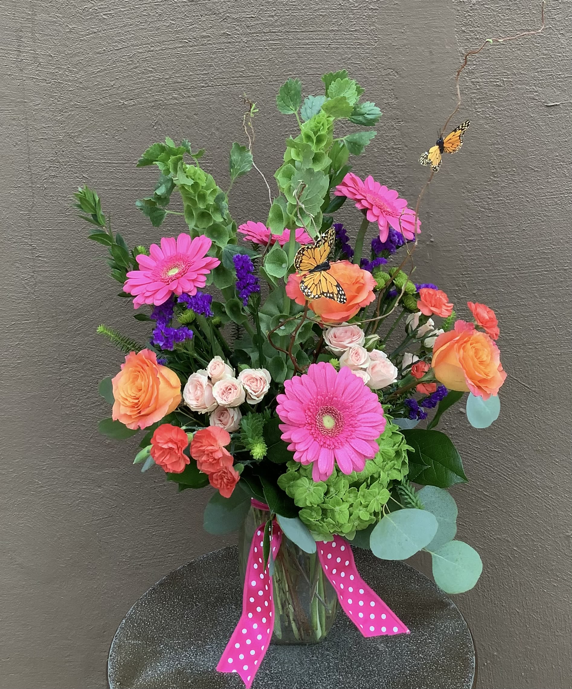 Butterfly Garden - Bright pink gerberas, orange and pale spray roses, coral carnations and purple fillers in a tall, garden-style vase with eucalyptus, butterfly picks and a pink polka-dot ribbon-cheerful gift-ready bloom.