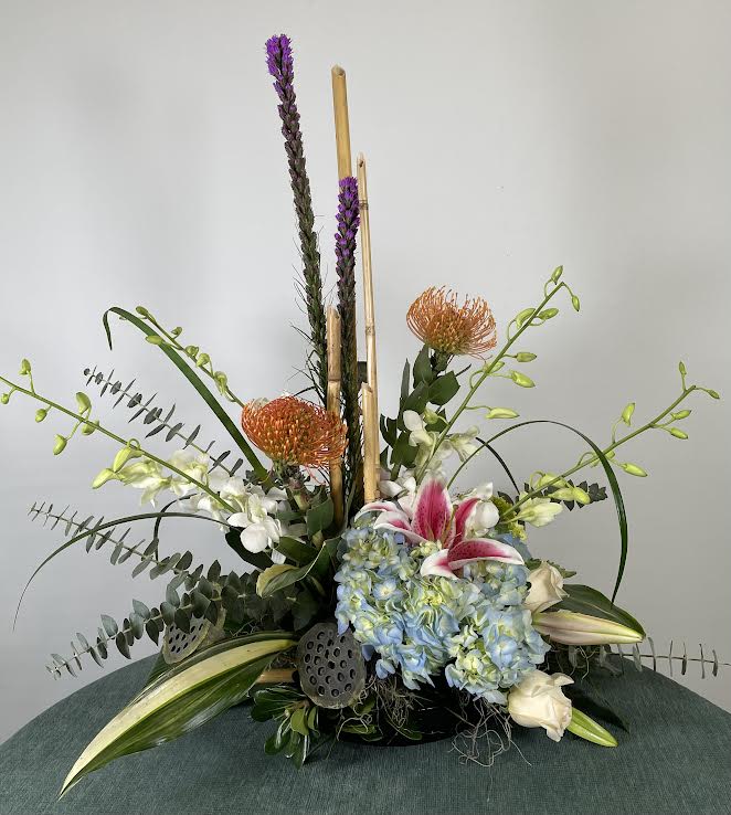 Tall floral arrangement with blue hydrangeas, lilies, and orange pincushion blooms