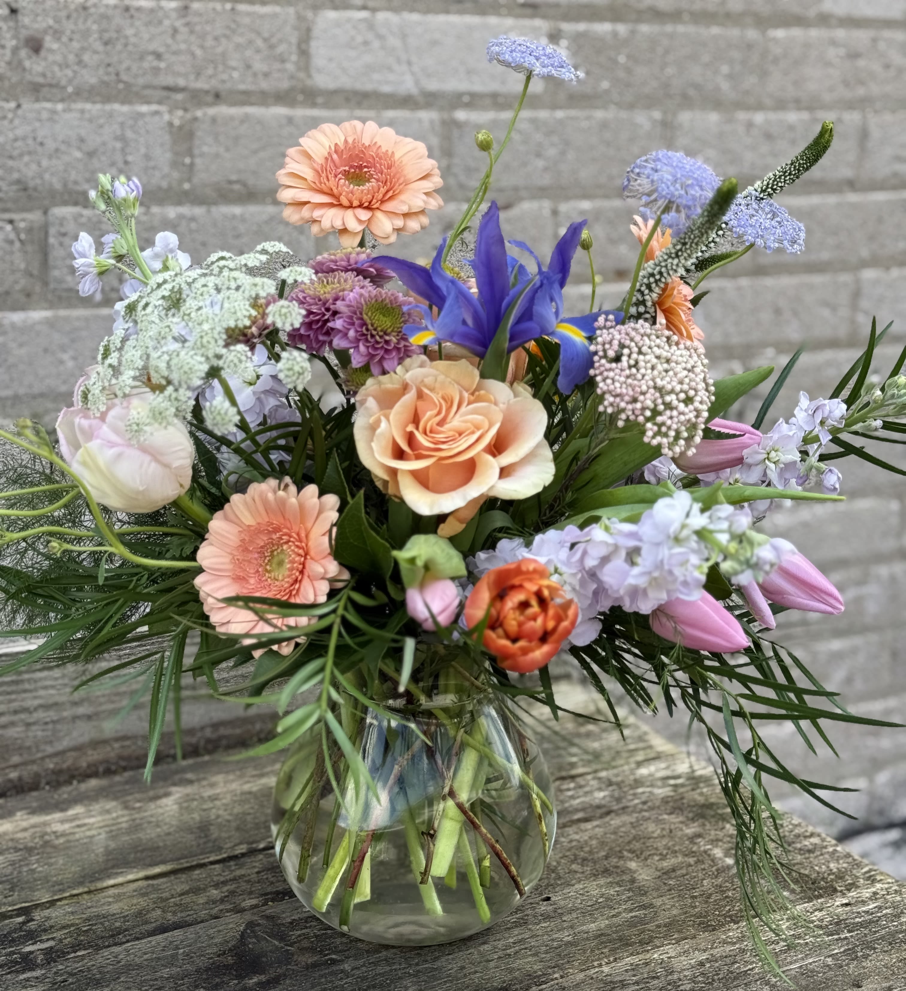 Seasons Appreciation - Peach gerbera daisies mingle with soft roses, pale pink tulips and deep purple iris in this Mother's Appreciation flower arrangement - a cheerful mixed-color bouquet perfect for Mother's Day, a birthday, or a just-because thank-you. The loose, garden-style gathering presents an airy, hand-tied look that reads fresh and joyful, the kind of arrangement that makes the person you're thinking of feel seen and celebrated.  Finished in a clear glass vase, the design pairs textured filler and clusters of small lavender blooms with chrysanthemums and other seasonal stems for contrast and movement. Colors range from warm peach and pink to cool lavender and purple, creating a balanced, springlike palette. Approximately vase-ready and sized for tabletops or counters; refresh water daily and trim stems every few days to extend vase life.