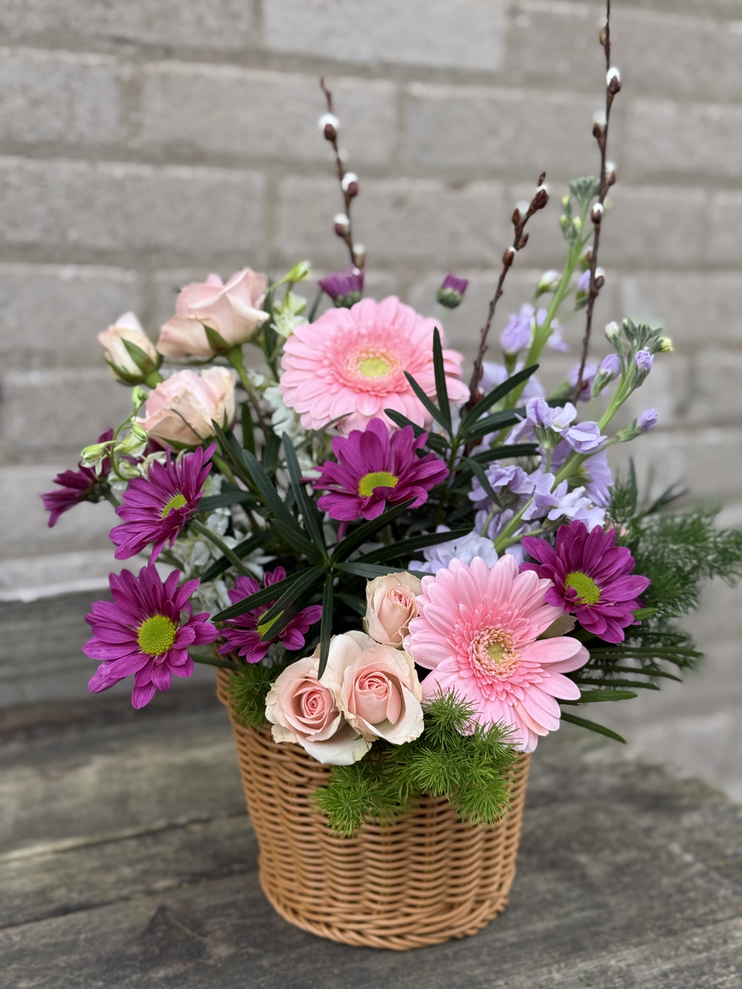 Summer Basket - Soft pink gerbera daisies and pale blush roses mingle with vivid purple daisy-like blooms in a woven wicker basket-an airy, garden-style flower arrangement perfect as a Mother's Day bouquet or a spring birthday gift. The tall branching stems and a mix of open blooms and buds give the composition height and movement so the person you're thinking of will feel genuinely celebrated the moment they see it.  Colors of pink, purple, white, and fresh green foliage create a feminine, cheerful mood that also suits occasions from anniversaries and get-well wishes to housewarmings, graduations, or a simple thank-you. Textures range from the smooth rose petals to the bold gerbera centers and daisy-like accents, arranged loosely for a natural, just-picked look. Presented in a wicker basket, it's easy to display and brings a casual, seasonal charm that works year-round-spring and summer especially-while making a thoughtful choice for Mother's Day, birthdays, sympathy, or "just because."