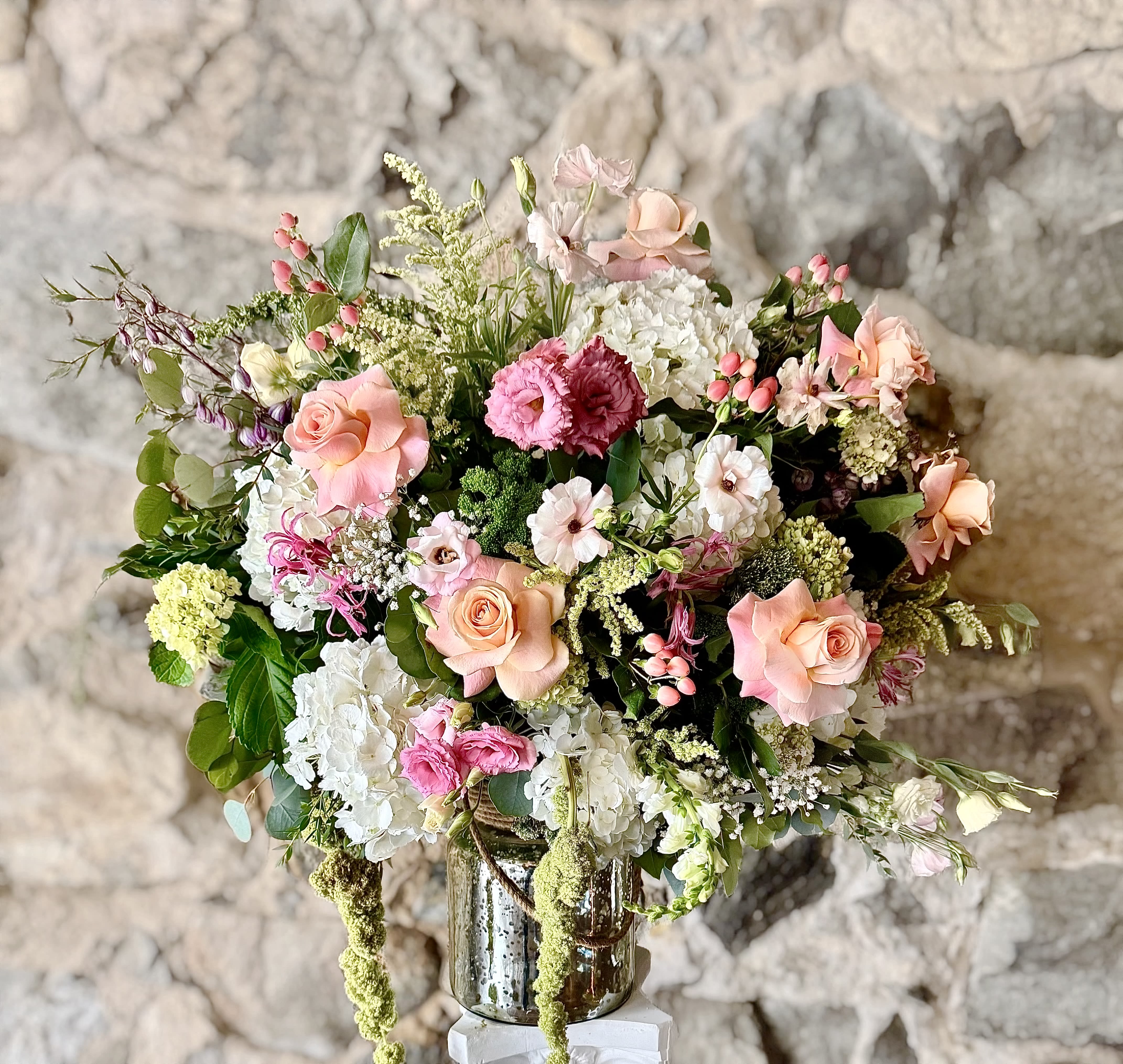 Fluer de Luxe - A soft, garden-fresh bouquet that reads royalty.  The Flue de Luxe arrangement greets the recipient with a full, rounded silhouette of pink, peach, white and green - the kind of floral surprise that makes someone feel truly seen and cherished.  This design layers garden roses, voluminous hydrangeas, delicate lisianthus and fragrant dianthus with smaller accent blossoms and airy filler for a textured, loose garden-style look. The mix of soft pinks and peaches against crisp whites gives the composition a gentle, timeless feel; the glass vase lets stems catch the light, adding understated sparkle to tabletops and mantels.  With simple care-fresh water and a cool spot-will keep the blooms looking their best through the week.