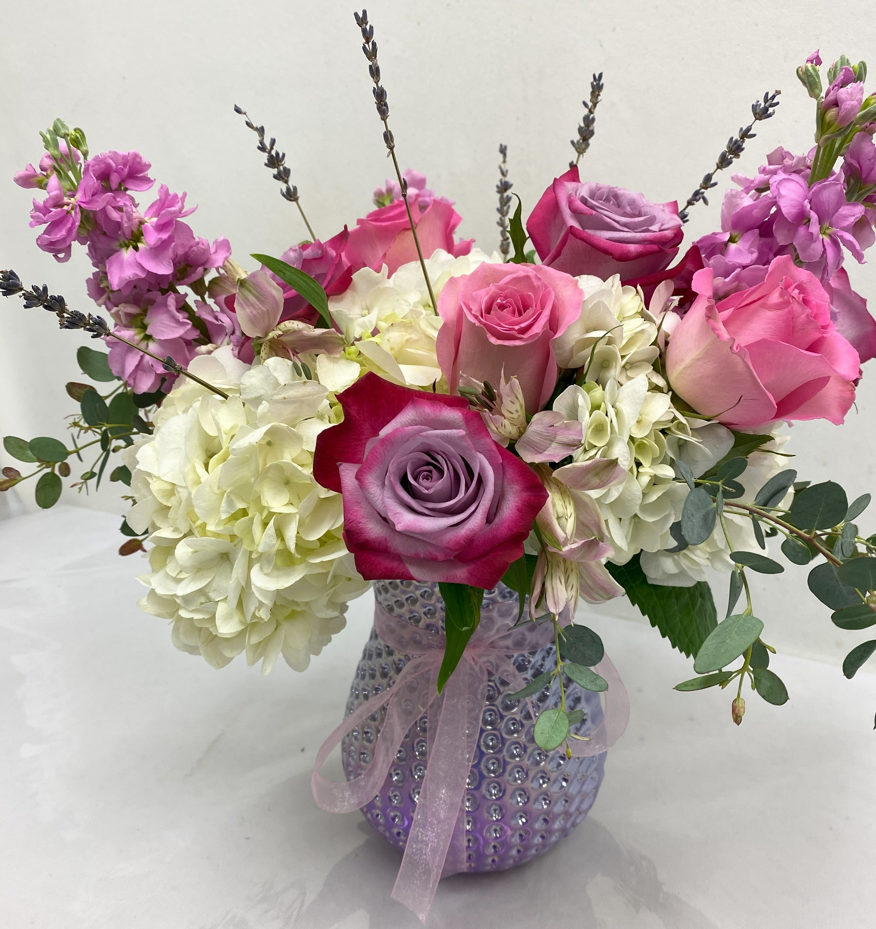 Pink and lavender roses with white hydrangeas in a textured lavender glass vase tied with ribbon
