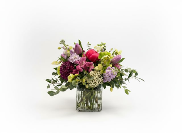 Luna - A lively, garden-inspired flower arrangement that feels like a breath of spring-perfect as a birthday bouquet, thank-you gift, or cheerful get-well surprise. This everyday floral arrangement places tulips and lily-like blooms center stage, paired with chrysanthemums, alstroemeria, and delicate filler stems to create a rounded, fresh silhouette that will make the person you're thinking of smile.  Soft pinks and purples mingle with crisp white and lime-green accents for a balanced, uplifting palette. The stems are arranged compactly with airy extensions for a relaxed, natural look and arrive in a clear square glass vase that showcases the clean lines and bright colors. Sized to sit nicely on a table or desk, Luna offers an elegant yet approachable presence that reads as thoughtful and joyful without being over-the-top.