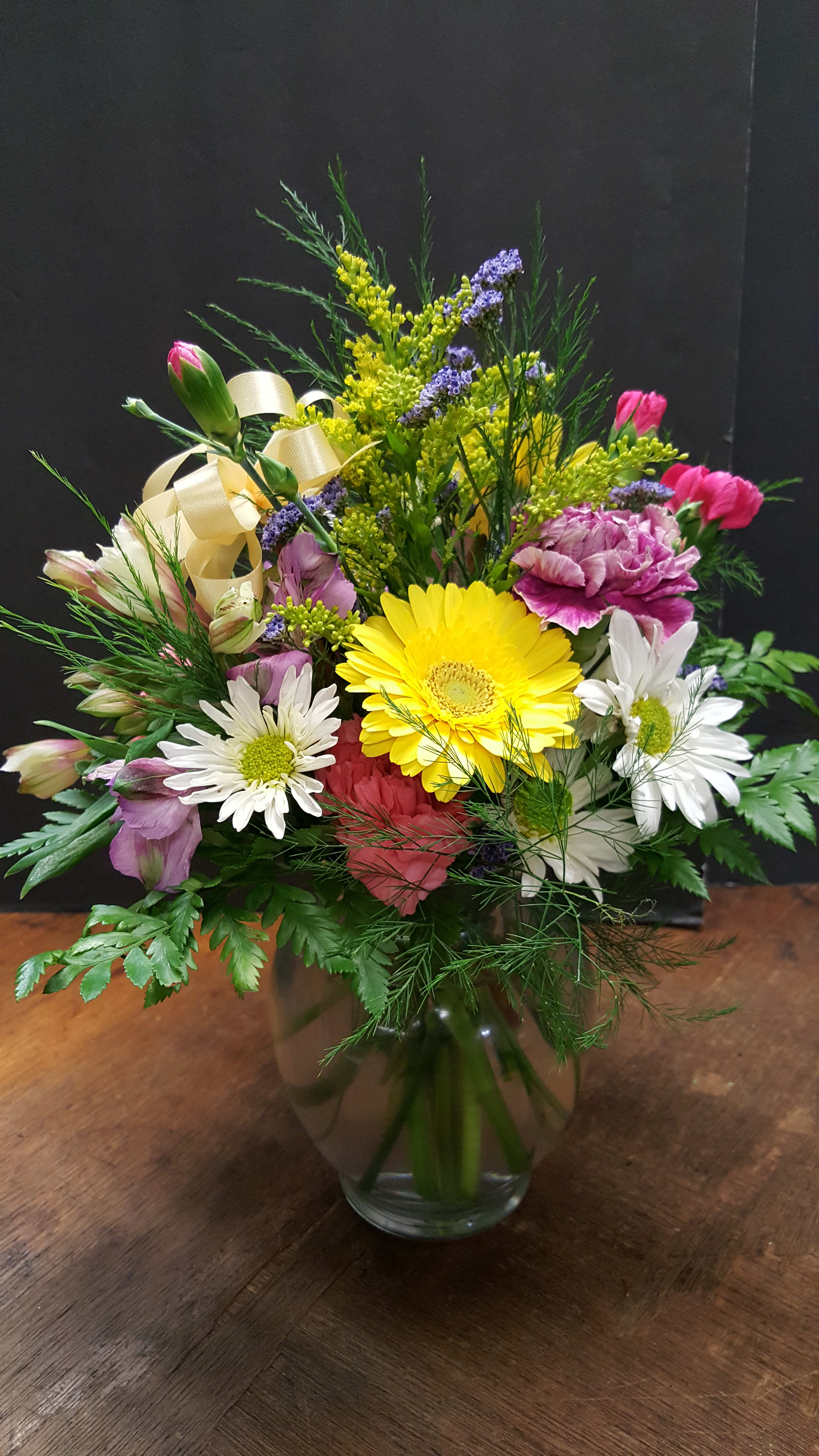 Mixed arrangement of yellow gerbera, white daisies, and pink carnations in a clear glass vase with ribbon.