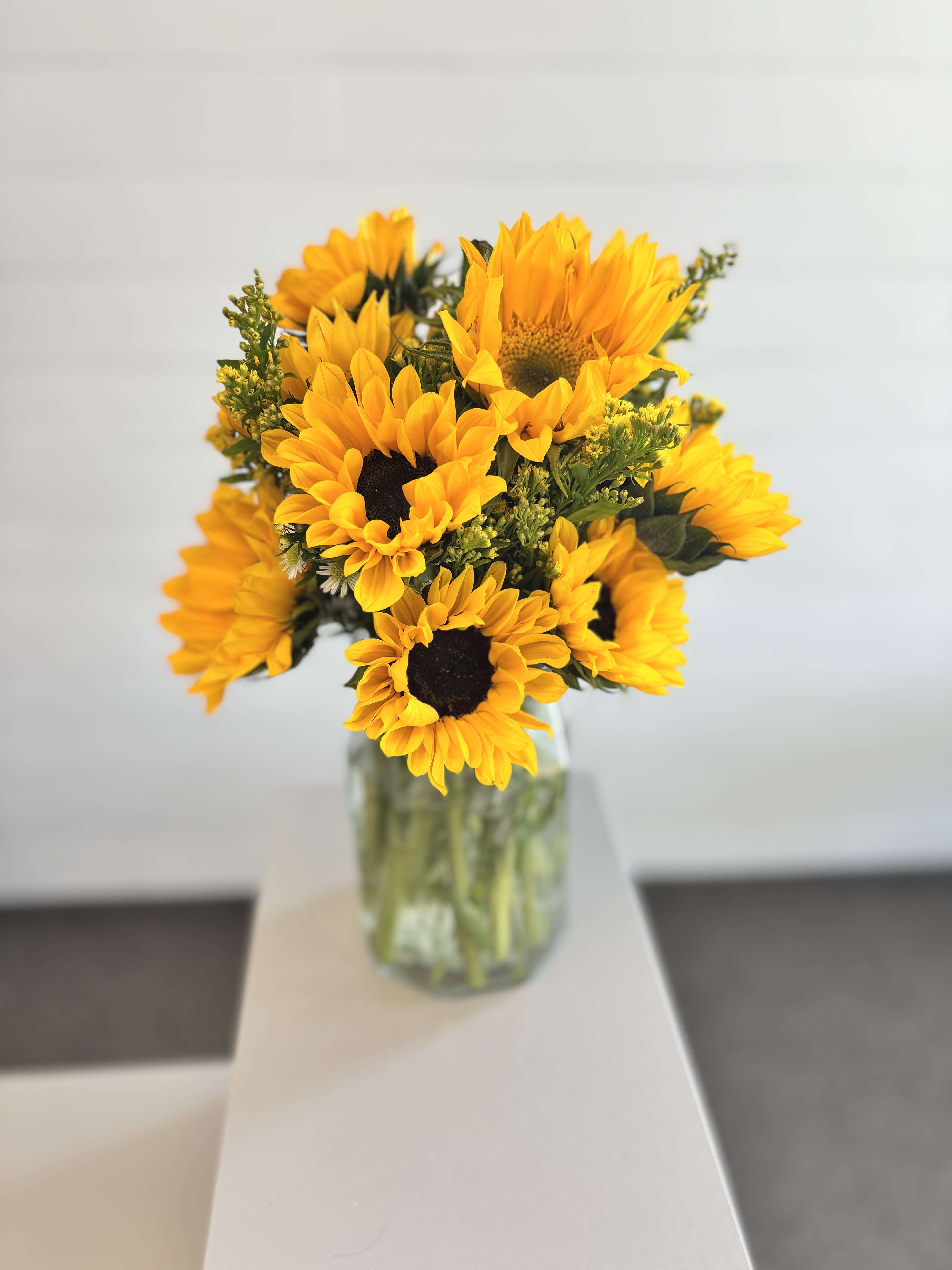 The Karima - Sunflower Vase Arrangement - A simple, radiant sunflower arrangement designed to bring warmth and light to any space. Clean and timeless, this design highlights the natural beauty of sunflowers, accented with fresh greenery and arranged in a clear glass vase.  Includes:  Fresh sunflowers Seasonal greenery Clear glass vase  Each arrangement is hand-designed, with slight variations based on bloom size and availability.