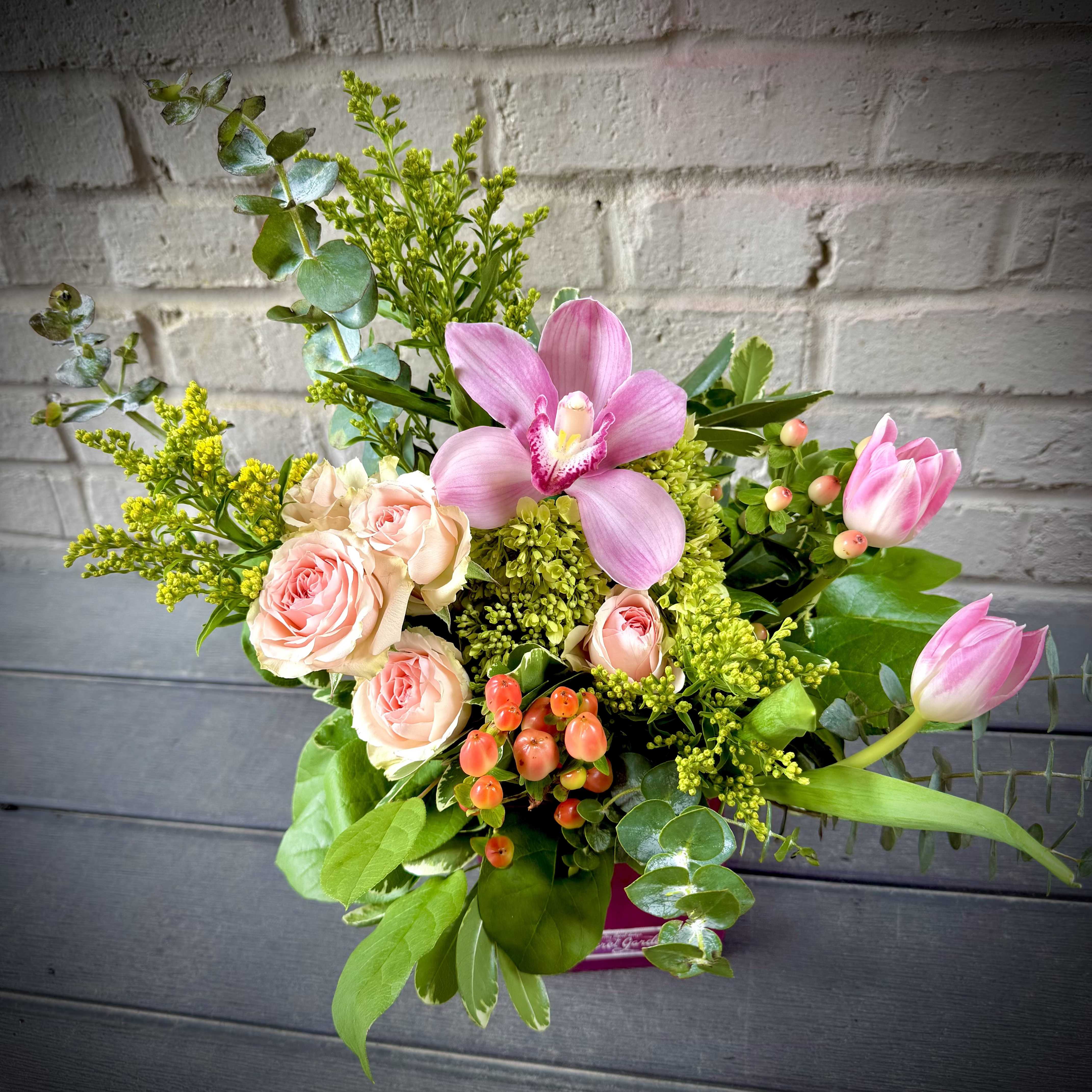Celebration Rose` - Soft pink orchids and blush roses take center stage in this celebratory flower arrangement, perfect as graduation flowers, a birthday bouquet, or a thoughtful "just because" gift. The magenta vase and pops of orange berries give the composition a lively, festive edge while the overall palette of pink, white, yellow, green and orange reads romantic and springlike-an ideal choice for marks of love and romance or to say you're thinking of them on a special day.  The design layers cymbidium and phalaenopsis orchids with spray roses, tulips, hydrangea accents, and airy yellow filler for textured, garden-style movement. Some stems reach upward and outward for a loose, asymmetrical silhouette while others form a full, rounded core; glossy green foliage and small berry clusters add contrast and depth. Choose the compact, layered presentation in a vivid magenta vase to suit the moment.  This arrangement feels joyful and celebratory for graduations, congratulations, and springtime occasions-she'll feel truly seen and celebrated when these blooms arrive. Stems are arranged for lasting shape; to keep flowers fresh, place in clean water and trim stems slightly before display.