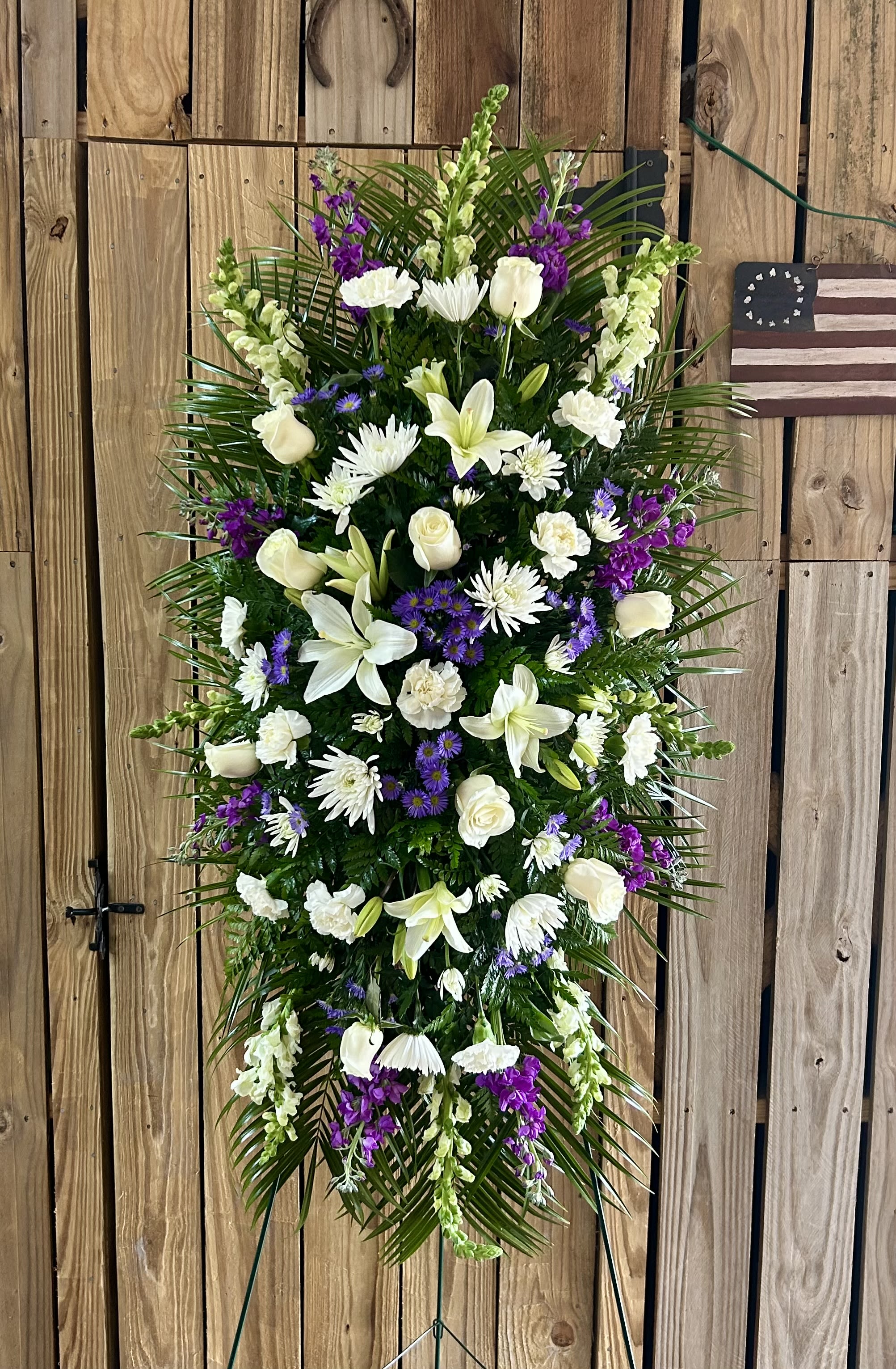 Majestic Memories Standing Spray - A tall, formal standing spray that reads with quiet dignity-this sympathy flower arrangement is designed to offer comfort at a funeral or memorial service. The Majestic Memories standing spray centers soft white lilies, roses, carnations, and daisy-like chrysanthemums, while vivid purple snapdragons and asters create solemn, graceful accents; the overall effect is elegant and respectful for a casket-side or easel display.  The spray is arranged on a traditional standing frame, layered with long palm fronds and lush greenery to create a full, upright silhouette that commands presence without showiness. Whites and deep purples are balanced across the composition so the lilies' trumpet shapes, the roses' layered petals, and the carnations' ruffled texture each read distinctly from a distance. This piece is designed to be tall enough for display on a stand at a service and to convey a message of sympathy and remembrance.  Choose this arrangement to honor someone who deserves a serene, dignified tribute-the kind of display that helps mourners feel seen and supported. Fresh-cut flowers arrive ready to place on a standing spray frame; for location-specific delivery or timing with a service, please contact us at checkout to coordinate.