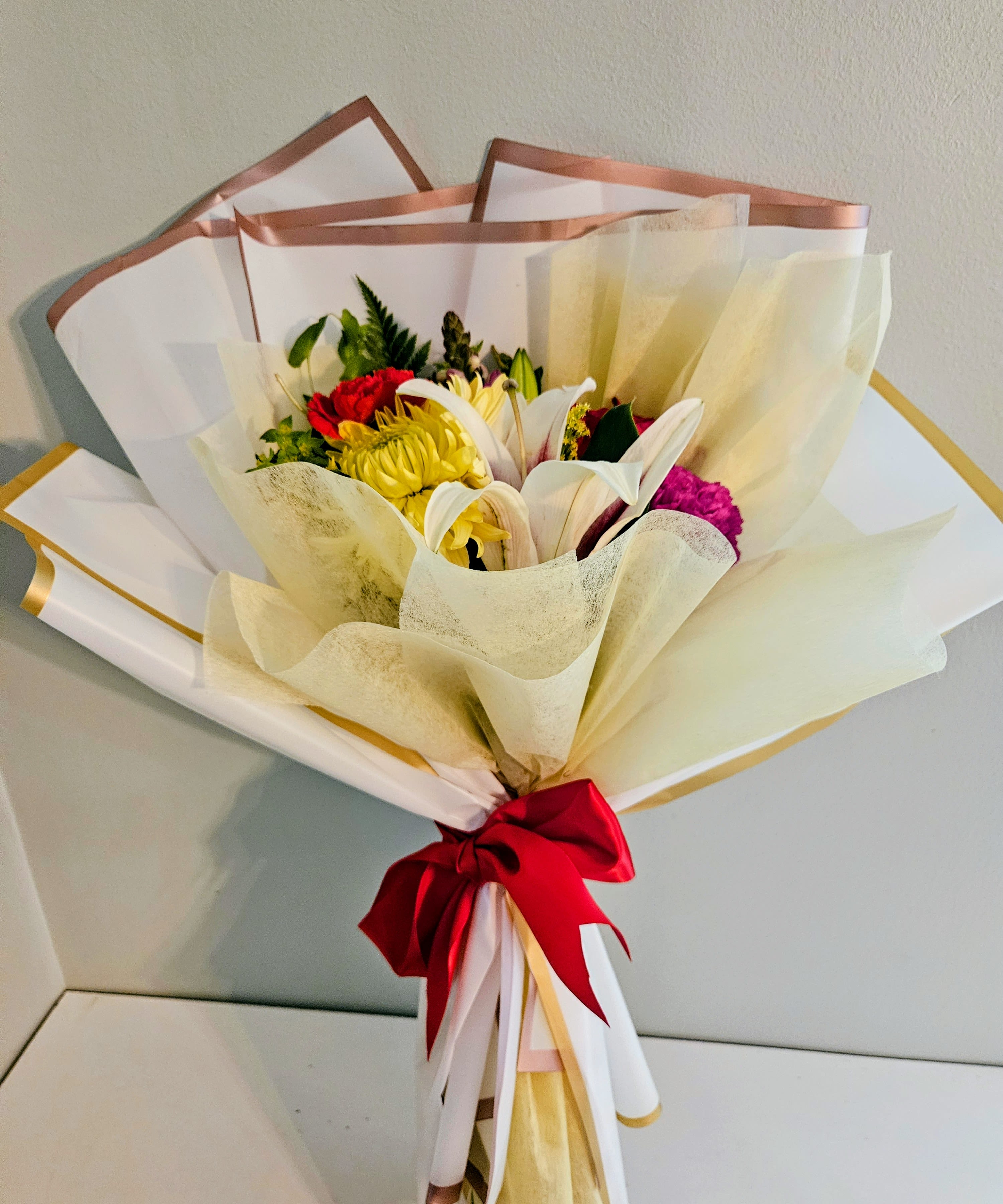 "Bright And Smile" - Sunlit yellow lilies and crisp white chrysanthemums meet cheerful pink carnations and classic red roses in a lively bouquet designed to make someone smile - ideal as a birthday bouquet, congratulations arrangement, or everyday thank-you gift. The mix of warm tones reads festive and upbeat, the kind of floral surprise that makes the recipient feel noticed and genuinely celebrated.  Wrapped in layered white and cream paper with metallic trim and finished with a large red satin ribbon, this arrangement arrives gift-ready with a polished, joyful presentation. The combination of lilies, chrysanthemums, carnations, and roses gives varied textures and lasting visual interest; stems are gathered securely at the base so the bouquet holds its shape and is easy to display.  Perfect for sending bright wishes to a friend, colleague, or loved one, this arrangement suits many occasions where you want to lift someone's spirits. For best longevity, trim stems and place in fresh water on arrival; if you'd like a vase, select one that comfortably fits the gathered stems to keep the display looking full.
