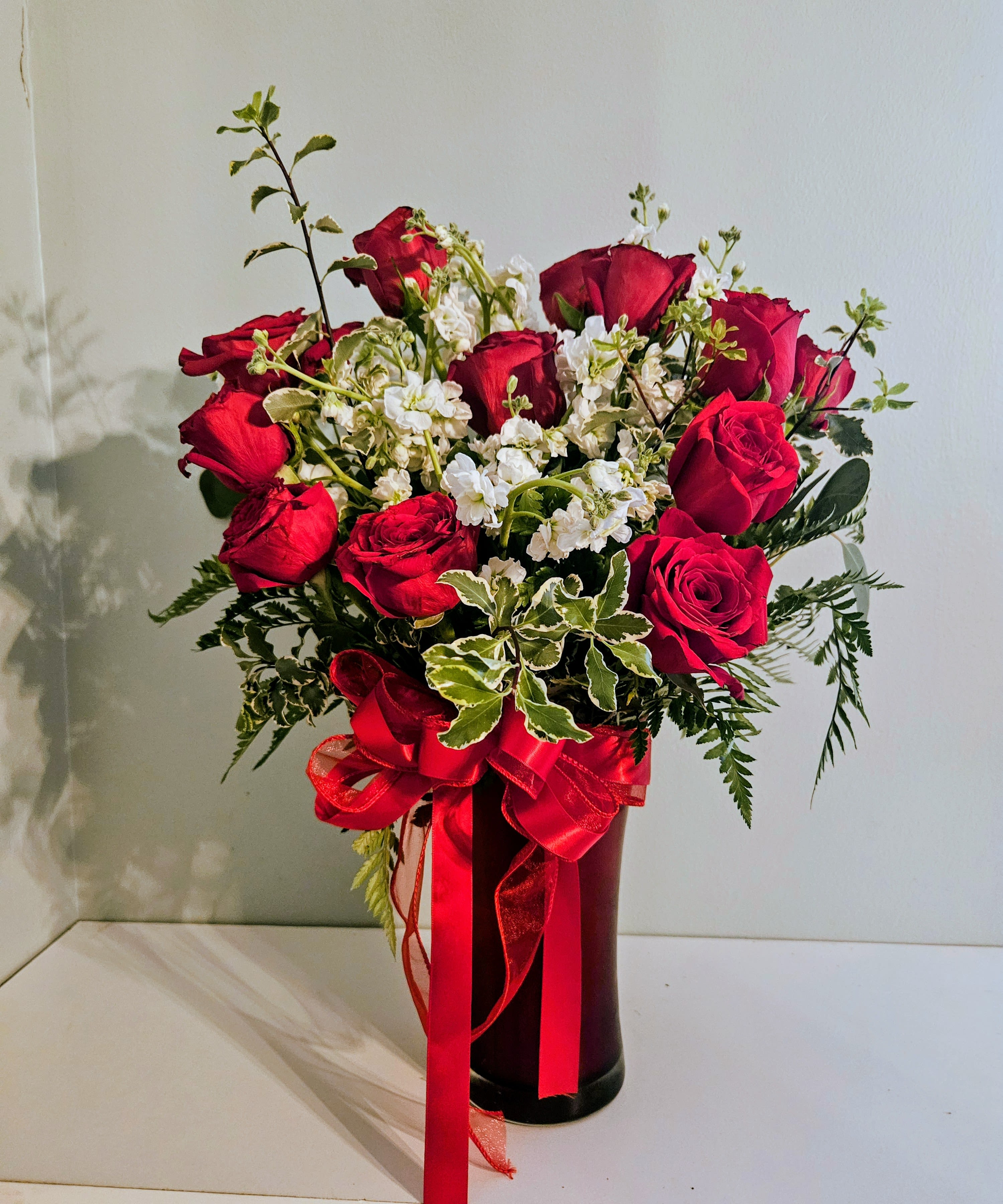 "Rose In Bloom " - A bold cluster of vivid red roses makes an immediate impression in this romantic flower arrangement-perfect as a birthday bouquet, anniversary roses gift, or a Valentine's Day surprise. The red-and-white contrast reads celebratory and elegant, so the person you're thinking of will feel cherished the moment they see it.  Dense red roses are accented with small white filler blooms and assorted greenery for texture and movement, gathered upright in a tall black vase and tied with a large red satin ribbon for a polished, classic finish. The design is full and centered, with blooms concentrated around the center and upper edges to create a strong visual mass that stands out on a table or mantle.  Ideal for expressing love, admiration, or congratulations, this arrangement suits romantic moments and festive everyday gifting alike. The tall vase gives the display presence and makes it easy to place on entryway consoles or dining tables; change the water every few days and trim stems if relocating to keep the roses looking fresh longer.