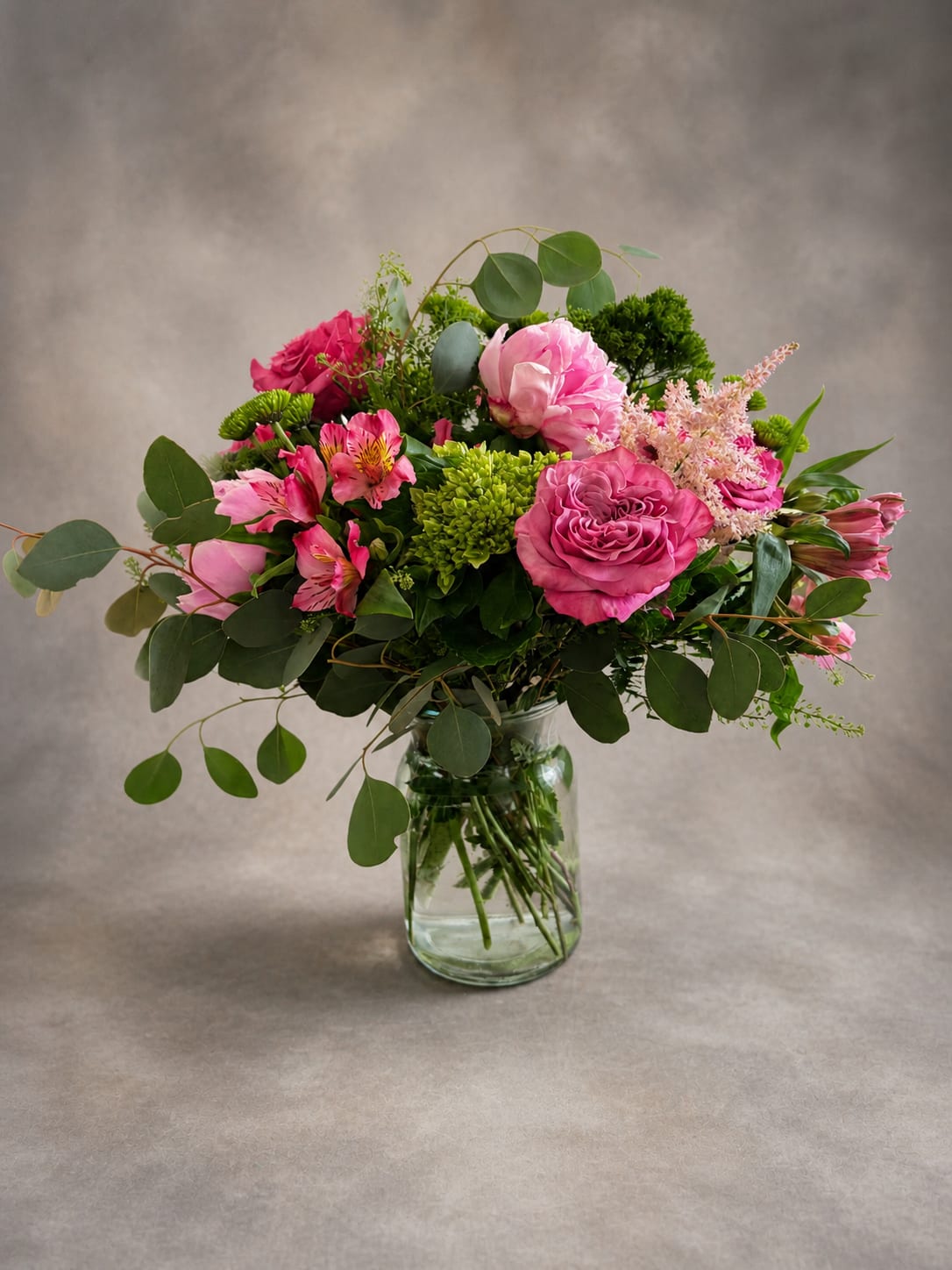 Pretty in pink -  Pretty in Pink is a lavish ode to all things feminine and flourishing. At its heart, full-bloomed hot pink garden roses and a blush peony unfurl with effortless grace, their layered petals rich with romance. Framing them, delicate alstroemeria in shades of magenta and blush dance alongside soft feathery astilbe, adding airy texture and a touch of whimsy. Clusters of chartreuse button mums bring a fresh, garden-market energy, while generous sweeps of silver-dollar eucalyptus and mixed greenery give the whole arrangement a wild, just-picked-from-the-garden soul. Nestled in a clear glass mason jar, Pretty in Pink feels as natural as it is stunning — the kind of bouquet that brightens a kitchen table, a bedside, or anywhere Mom deserves a little extra beauty. It's the perfect way to say thank you to the woman whose love is as full and generous as the flowers themselves.