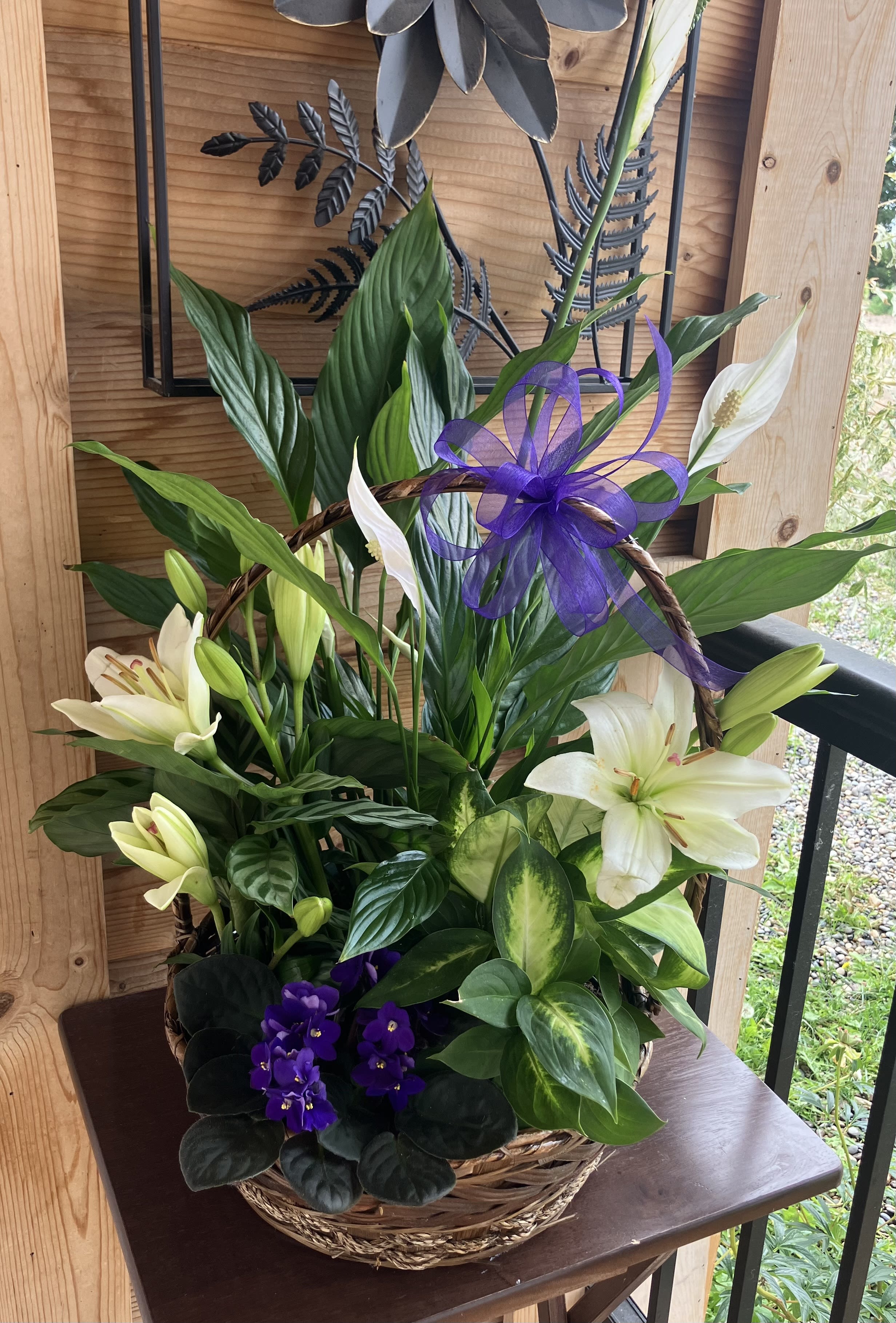 Basket of mixed green plants with white lilies and purple flowers, topped with a purple ribbon bow.