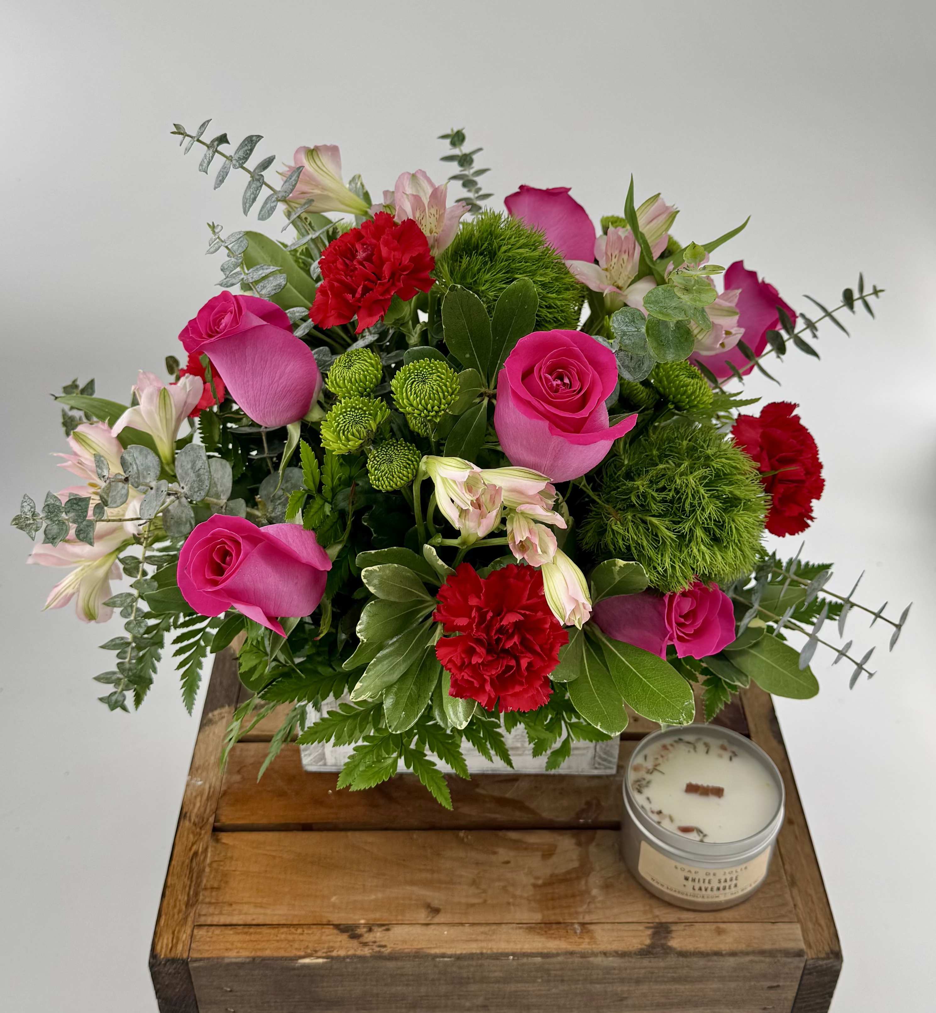 Arrangement of hot pink roses, red carnations, and green accents in a white box with a small candle beside it.