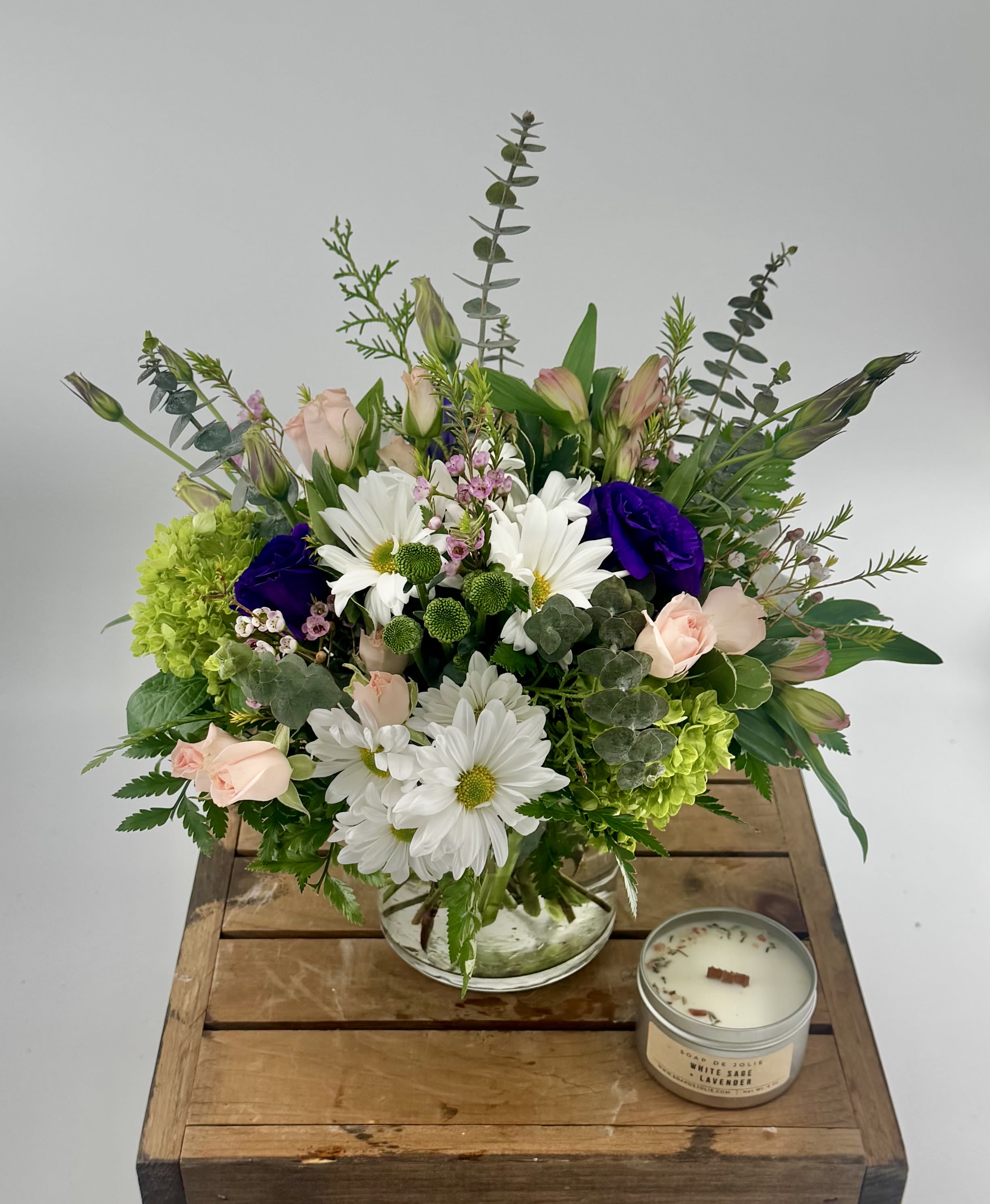 Mixed bouquet of white daisies, pink roses, and purple blooms in a glass bowl with a candle on a wooden crate
