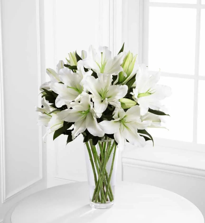 Tall bouquet of white lilies with green buds in a clear glass vase on a white table
