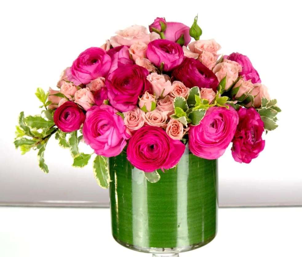 Pink ranunculus and pale pink spray roses in a leaf-lined clear glass cylinder vase