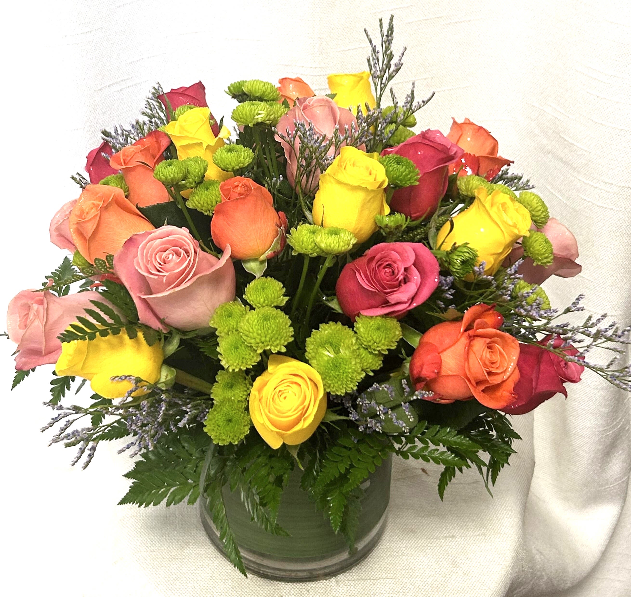 Colorful bouquet of mixed roses and green button mums arranged in a clear glass cylinder vase.