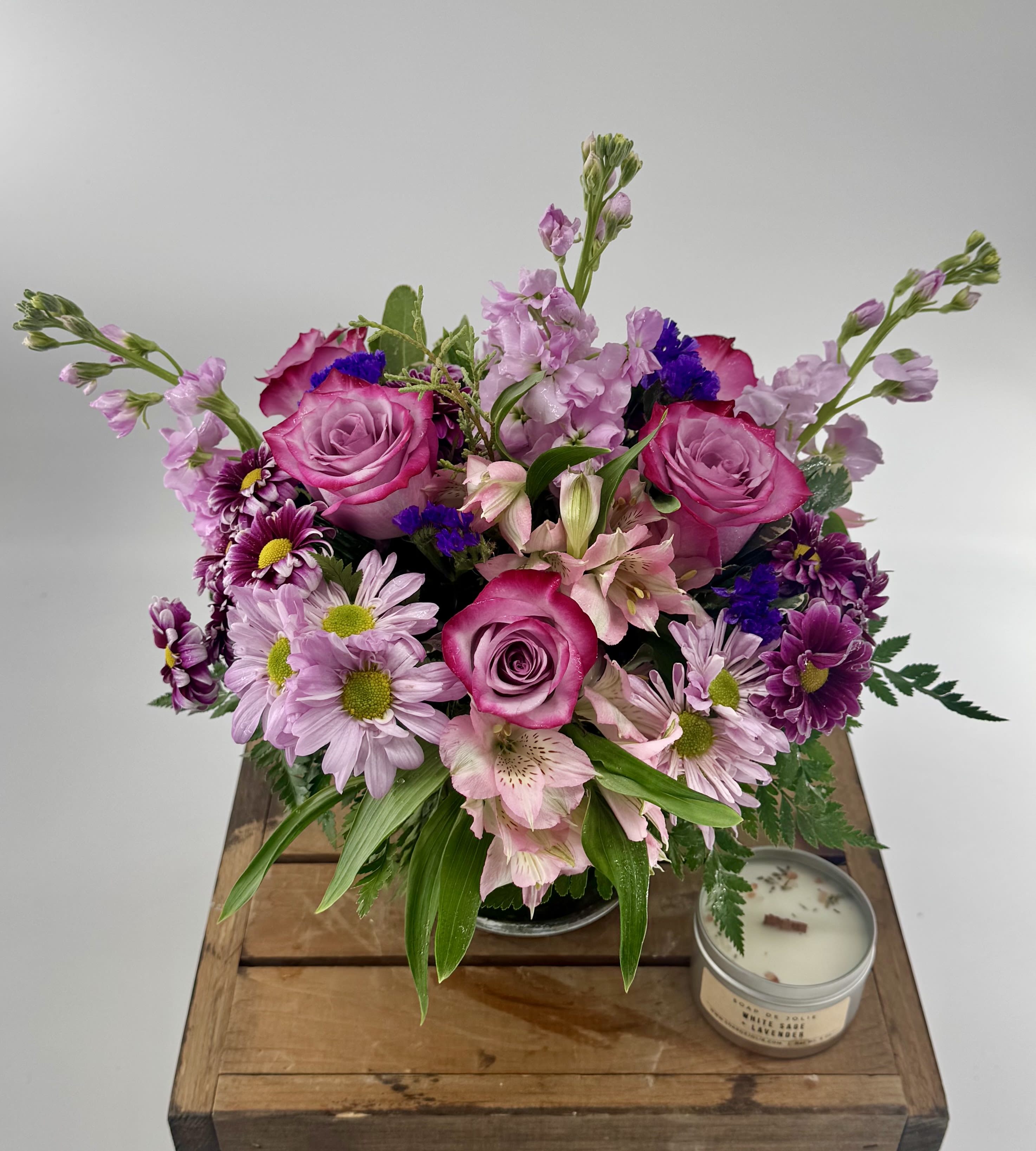 Round bouquet of pink roses, lavender mums, and alstroemeria in a glass vase beside a small candle.