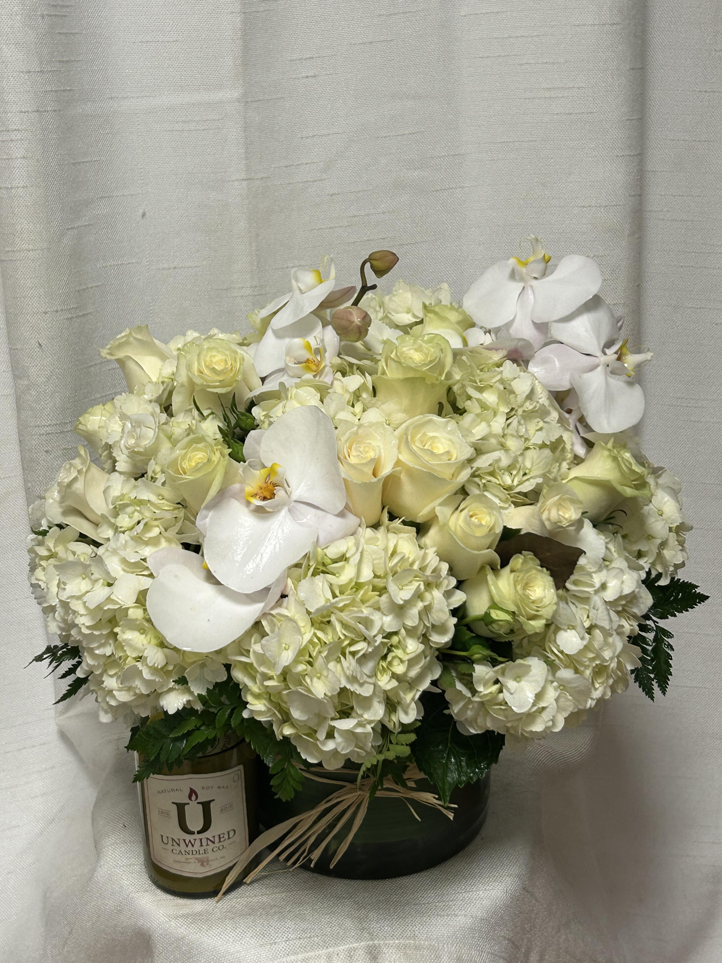 Low round arrangement of white roses, hydrangeas, and orchids in a green vase with a candle beside it