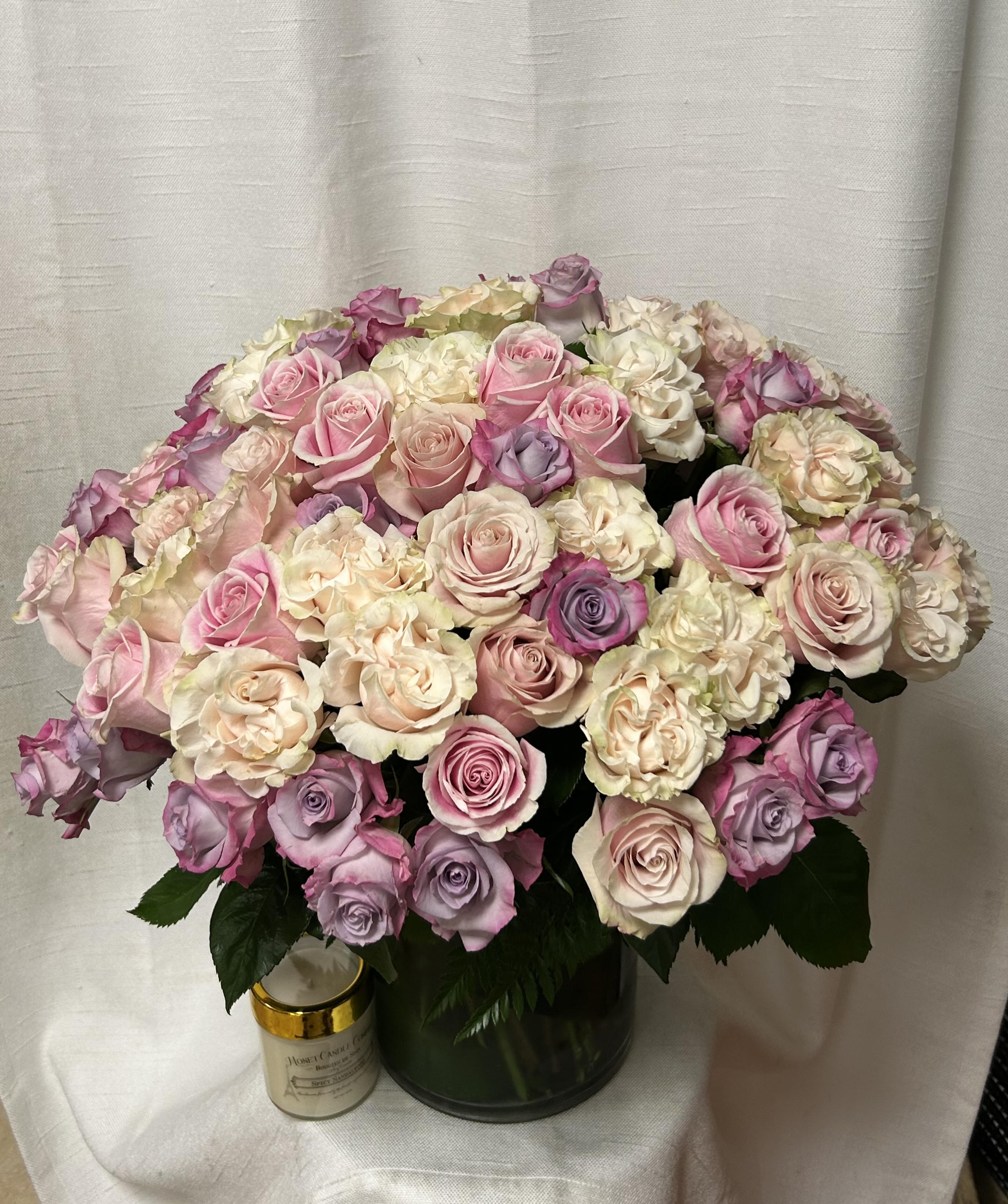 Large arrangement of pastel pink, cream, and lavender roses in a glass vase with a candle beside it