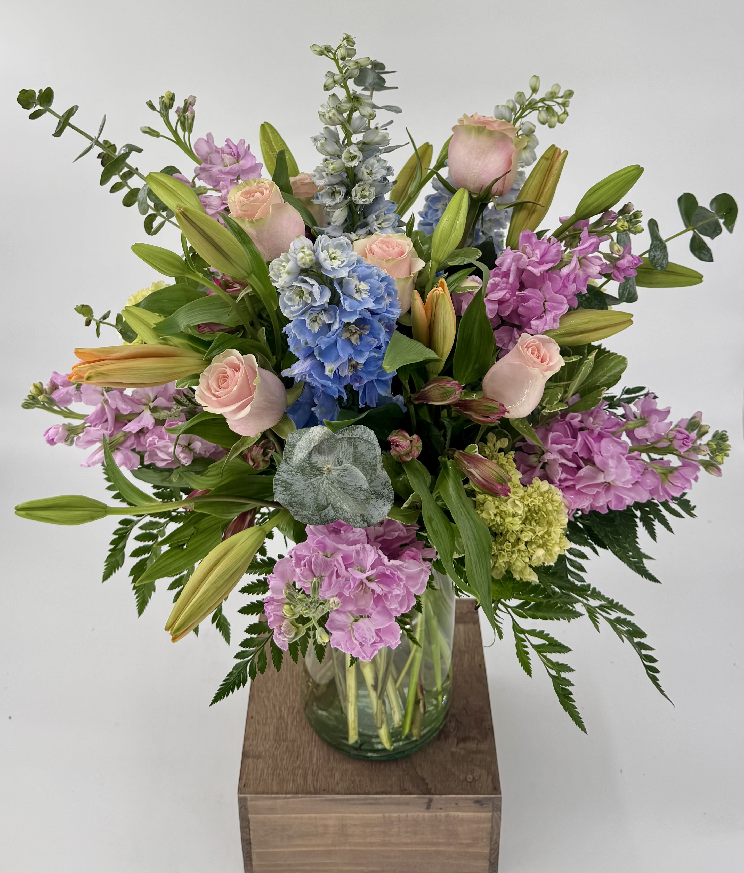 Tall pastel arrangement of pink roses, lilies, blue delphinium and stock in a clear glass vase
