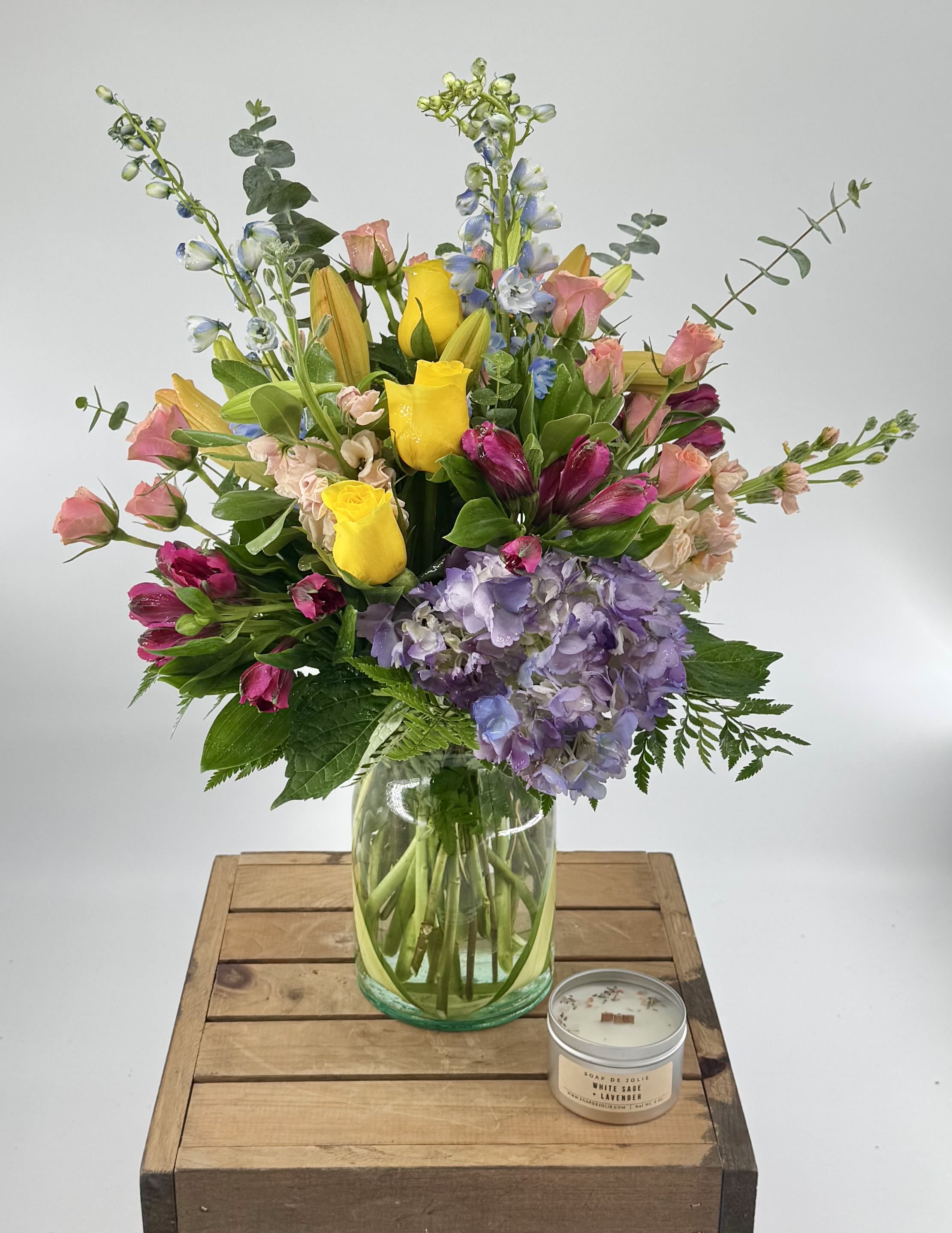 Tall mixed bouquet with yellow roses, lilies, hydrangea, and other flowers in a glass vase beside a small candle