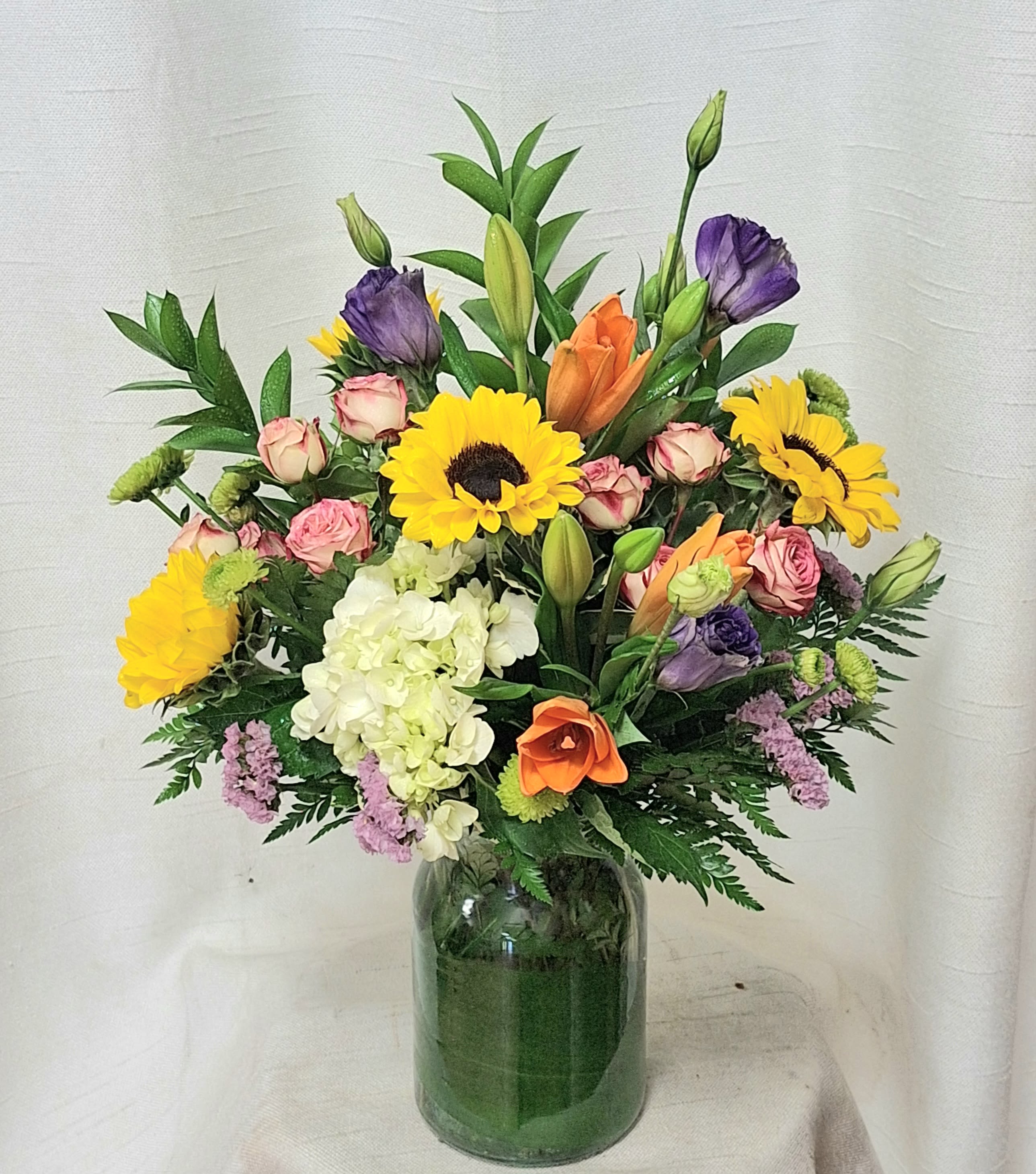 Mixed bouquet of bright flowers including sunflowers, lilies, and roses in a clear glass vase.