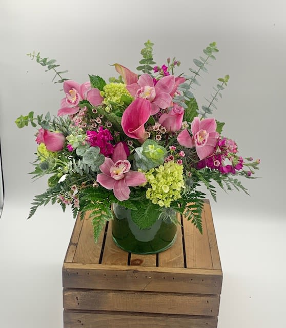 Pink calla lilies, orchids, and green hydrangeas in a clear glass vase on a wooden crate