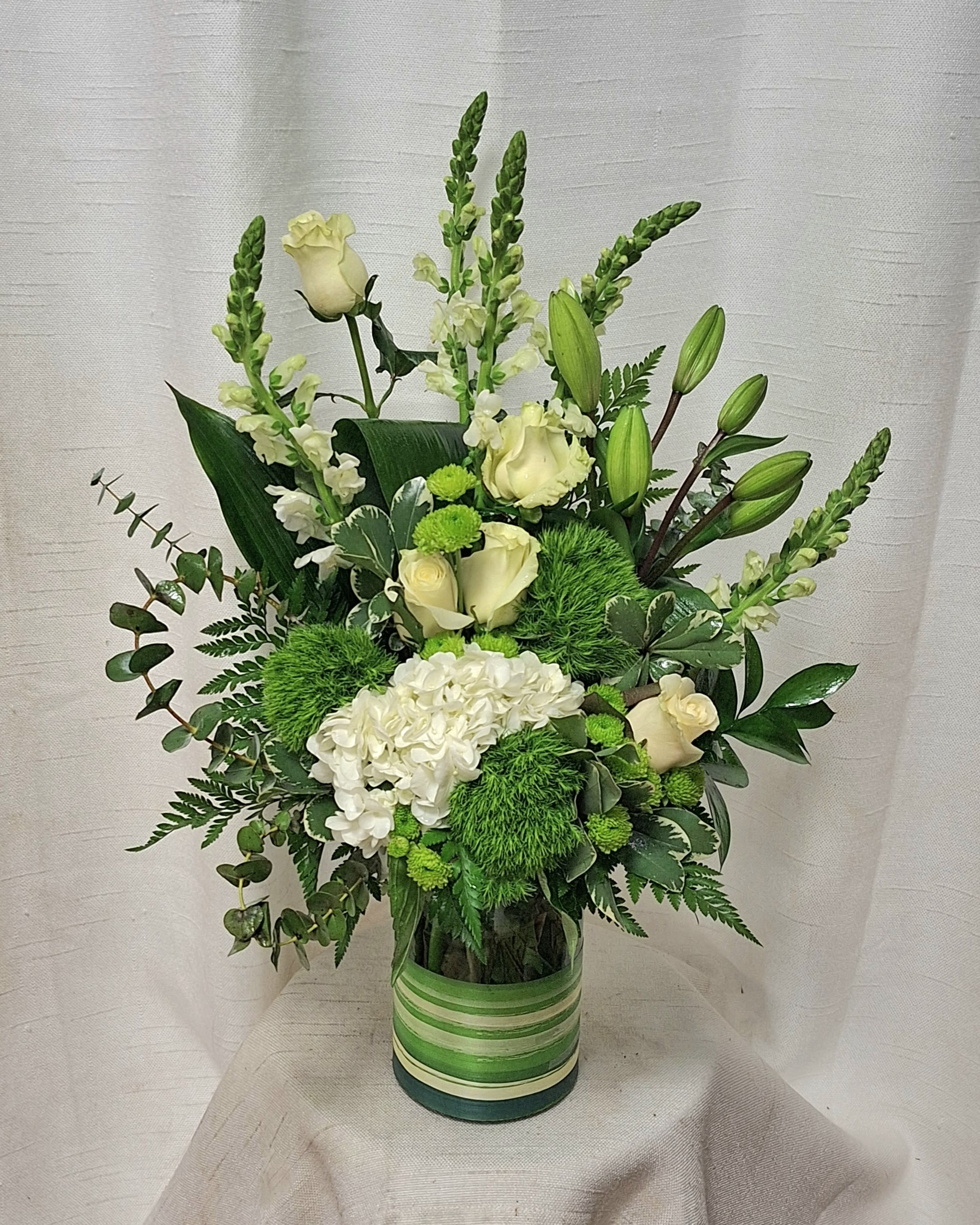 Botanic Delight (DL) - Step into a realm of pure sophistication and timeless beauty with our "Ethereal Elegance" floral arrangement, a graceful masterpiece that exudes a sense of tranquility and refined elegance. This enchanting composition features a sublime combination of pristine white roses, verdant green dianthus, delicate white lilies, majestic white snapdragons, and luxuriant premium greenery, coming together in a harmonious display of purity and poise.