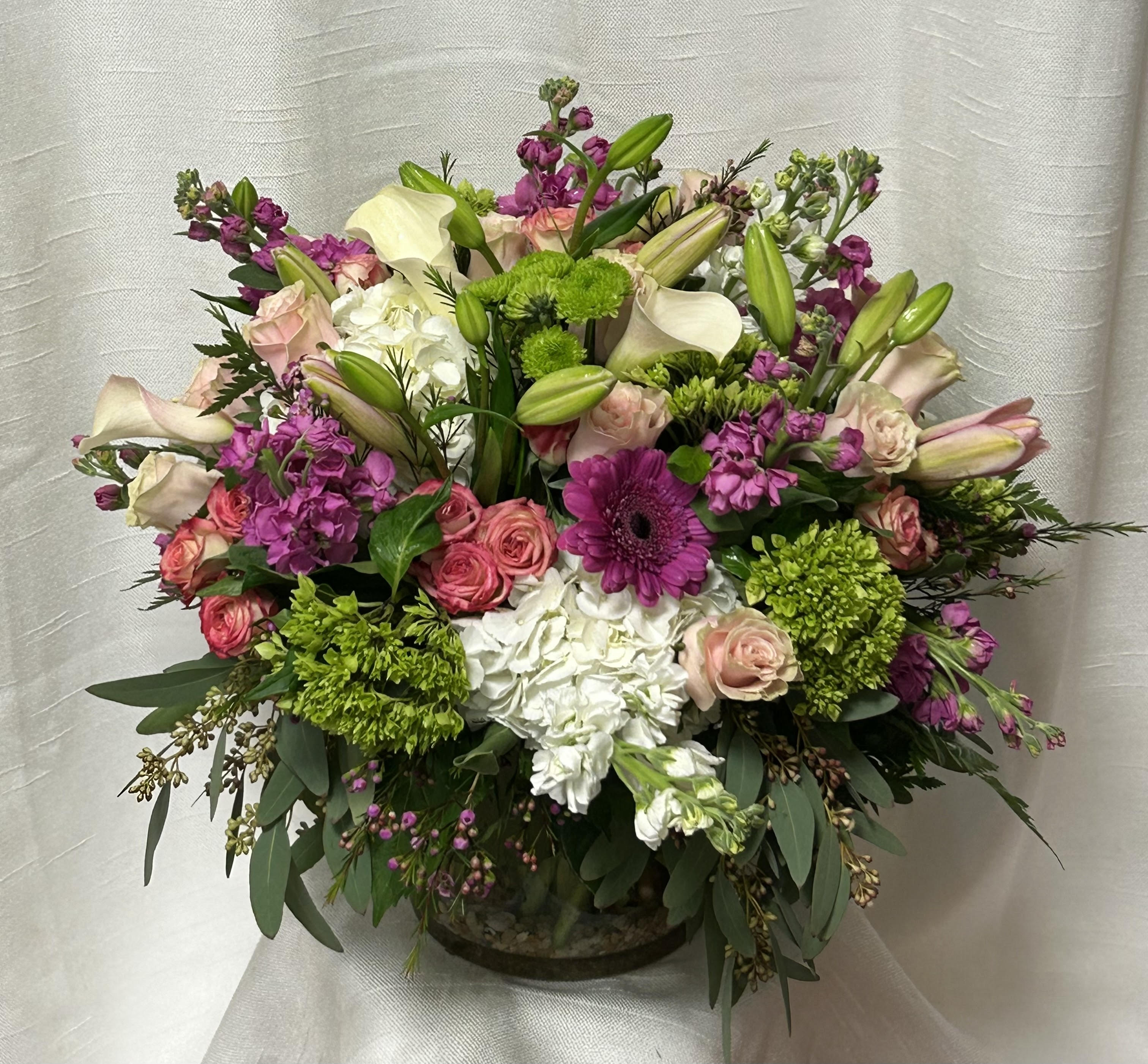 Premium Pastels (DL) - Experience the essence of modern sophistication with our exquisite modern floral arrangement. This stunning arrangement features a striking combination of green and white hydrangeas, mini callas, lilies, roses, spray roses, gerberas, wax flowers, stock, and more. Expertly designed in a large leaf-lined glass cylinder vase, this arrangement is the perfect example of contemporary elegance and style.  