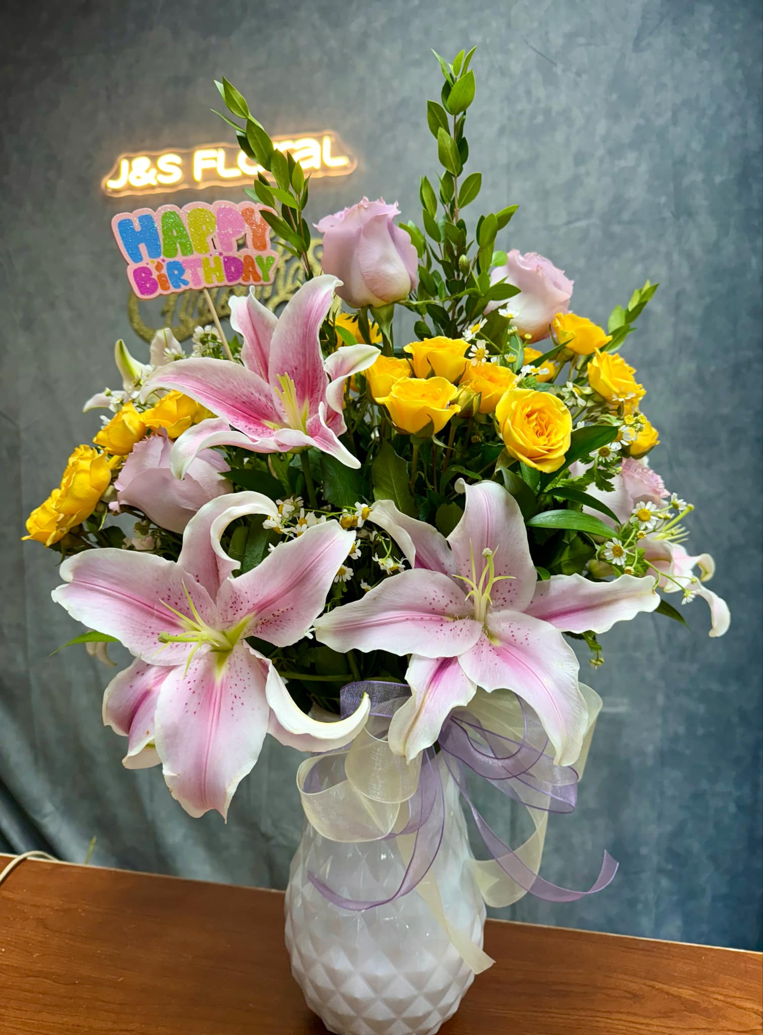 Pink lilies and yellow roses in a white vase with a birthday sign