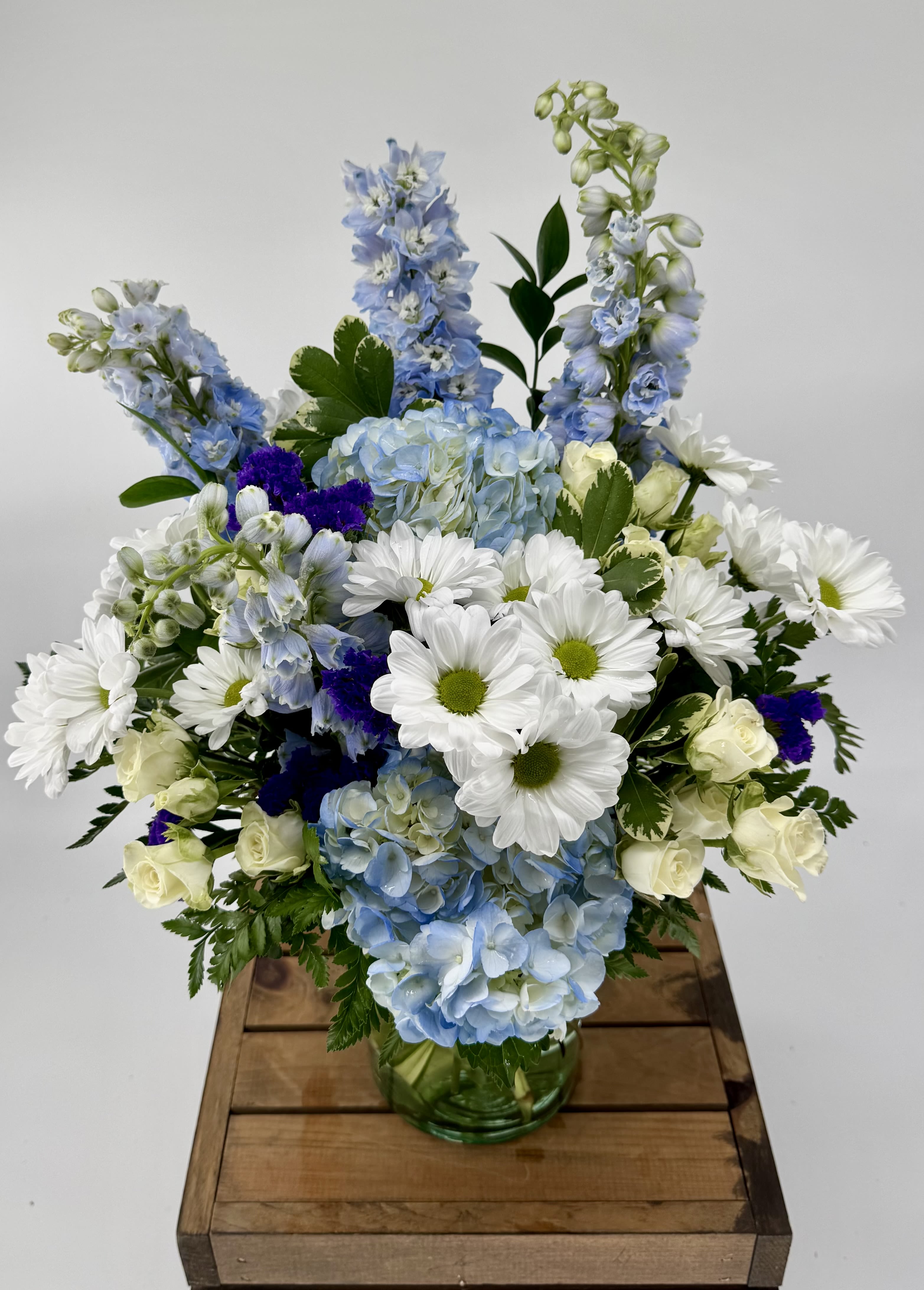Sky Blue (DL) - Immerse yourself in a tranquil oasis with our enchanting Sky-Blue flower arrangement, showcasing a harmonious blend of blue hydrangeas, white spray roses, blue delphiniums, purple statice, white daisies and assorted greens. Artfully arranged in a leaf-lined glass cylinder vase, this stunning ensemble evokes a sense of serenity and beauty.