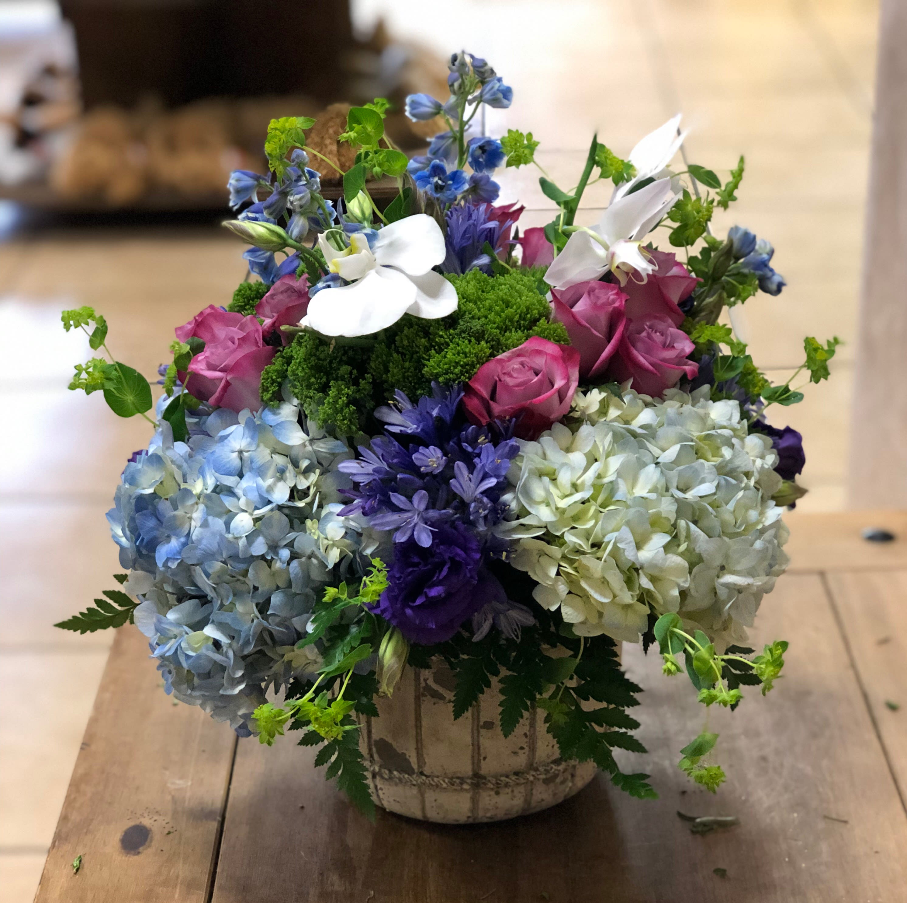 Moody Blues (BB) - Shades of Green, Blue and Purple in a decorative container, perfect for all occasions! Includes purple Roses, blue Hydrangea, blue Agapanthus, Orchids, Trachelium, Delphinium and more. Come artistically arranged in a decorative container.