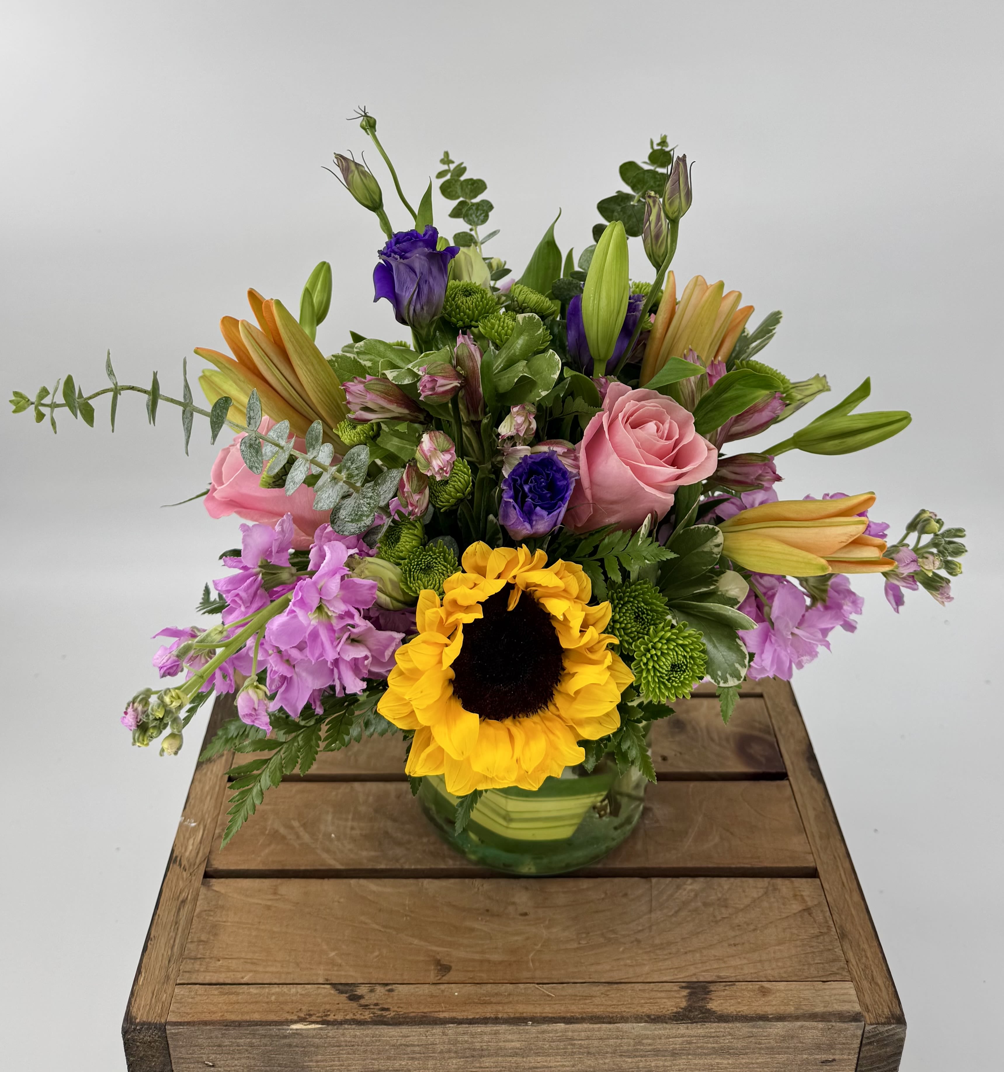  Radiant Joy (BB) - A vibrant and joyful arrangement that is sure to brighten anyone's day. This delightful bouquet features a lively combination of orange lilies, sunflowers, pink stock, purple lisianthus, green button poms, and pink roses.