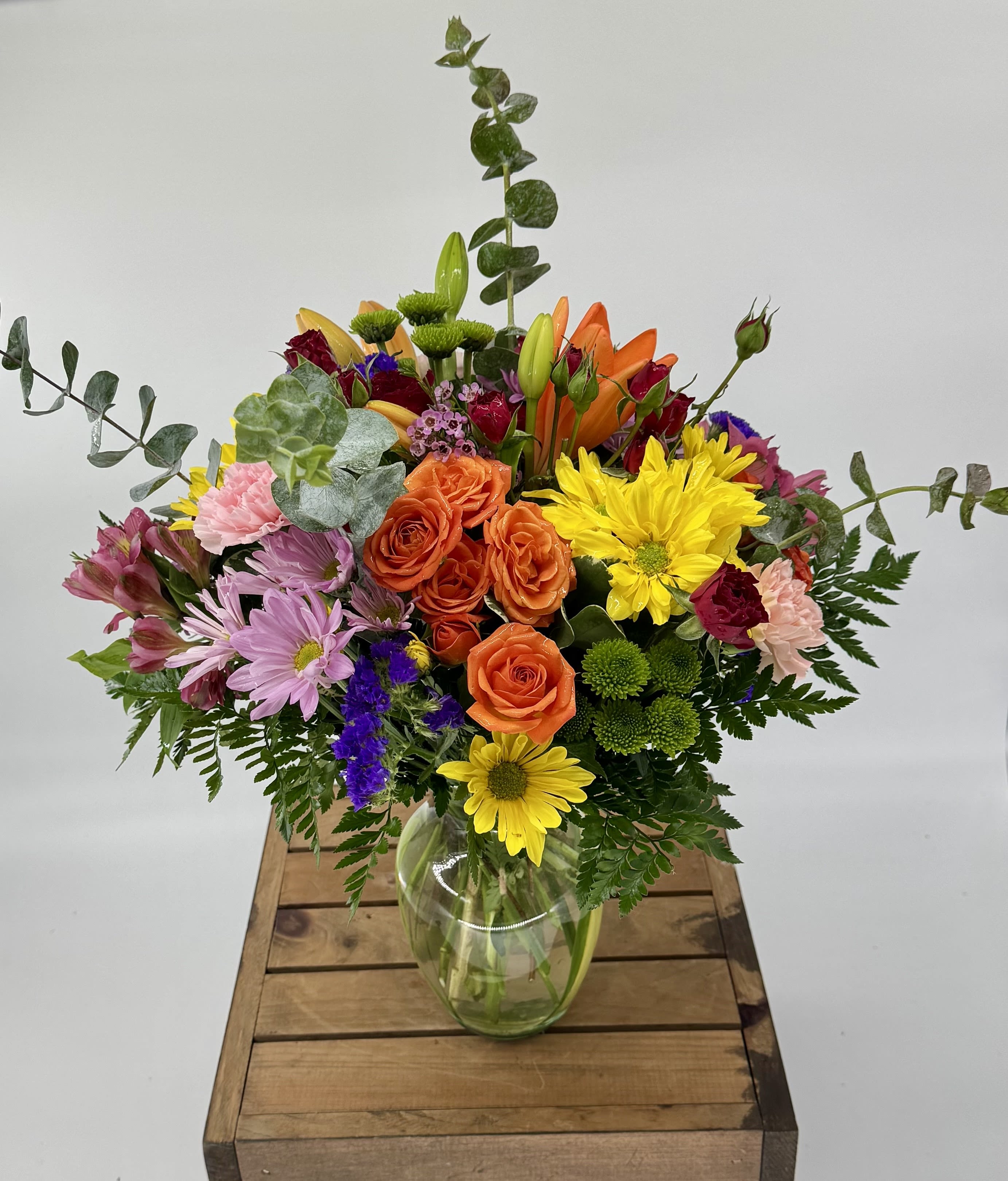 Color Cascade (DL) - This vibrant modern floral arrangement features orange lilies, yellow and lavender daisies, purple statice, red and orange spray roses, pink carnations, eucalyptus, waxflowers, and assorted greens, beautifully arranged in a clear glass vase for a fresh, colorful display. 