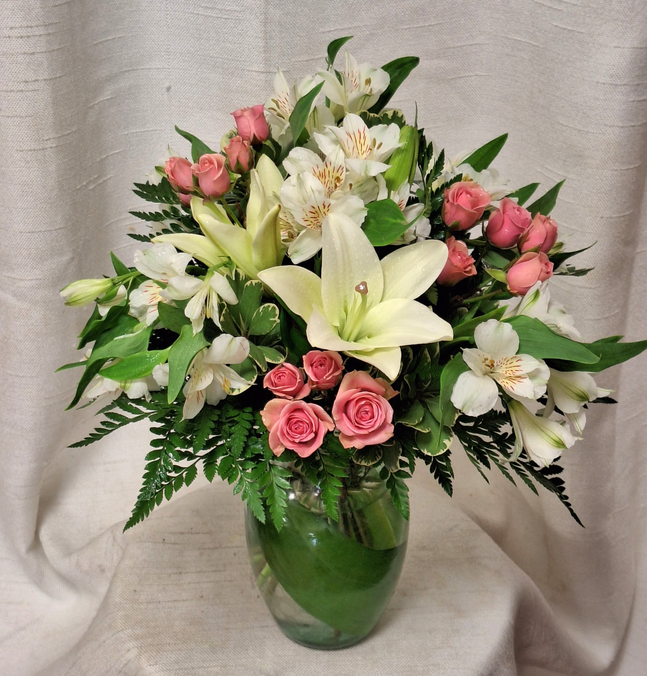 In Style (BB) - Introducing a stunning flower arrangement that exudes elegance and charm. This exquisite bouquet features a delicate blend of soft pink roses, white lilies, white daisies, and white alstroemeria, expertly arranged in a glass vase with lush greenery and filler for a touch of natural beauty.