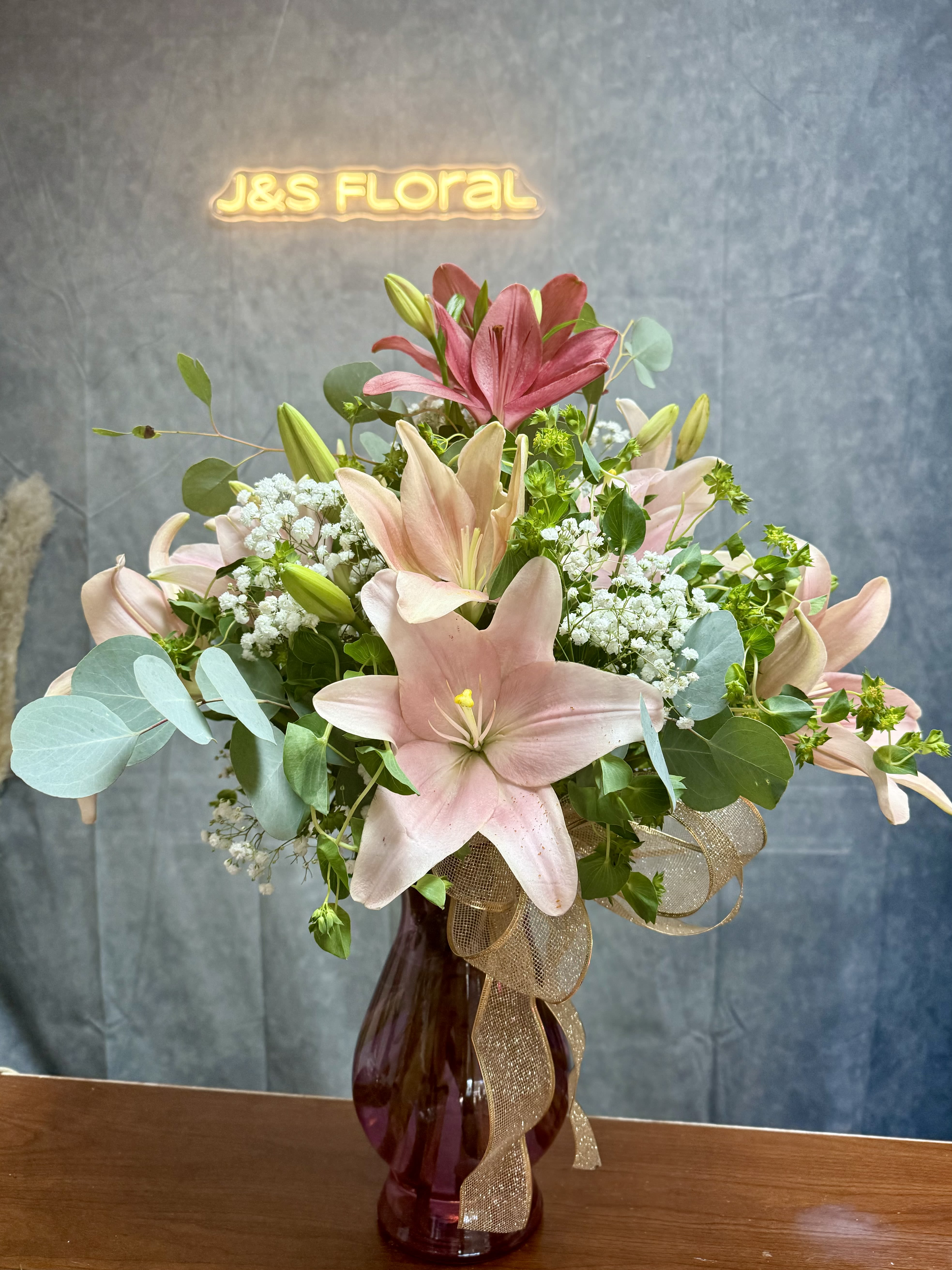 Pink lilies and baby's breath in a burgundy vase with a gold ribbon