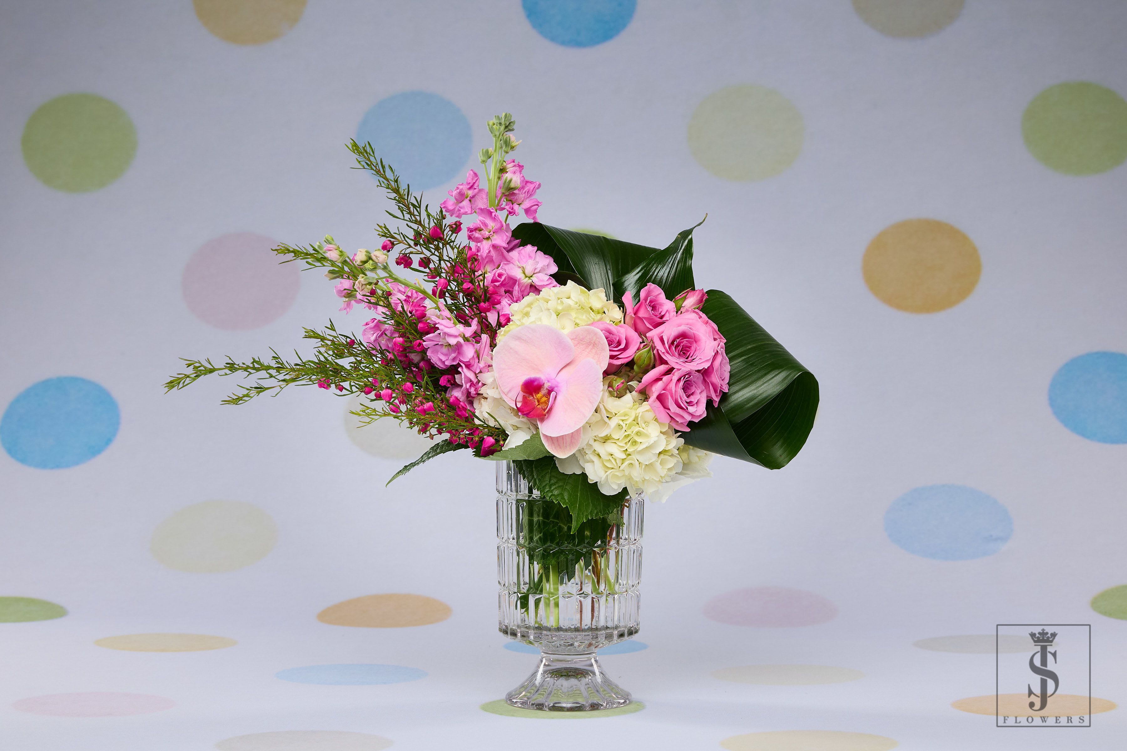 Blushing Elegance - Blushing Elegance is a stunning symphony of romantic tones and refined texture. Featuring a lush mix of vibrant pink roses, stock flowers, and a show-stopping orchid bloom, this arrangement is grounded in soft white hydrangeas and accented with airy waxflowers. Nestled in a vintage-style cut crystal vase, it’s both timeless and modern — ideal for upscale gifting, anniversaries, or simply to make someone feel deeply appreciated.  This piece radiates sophistication, femininity, and heartfelt beauty.  
