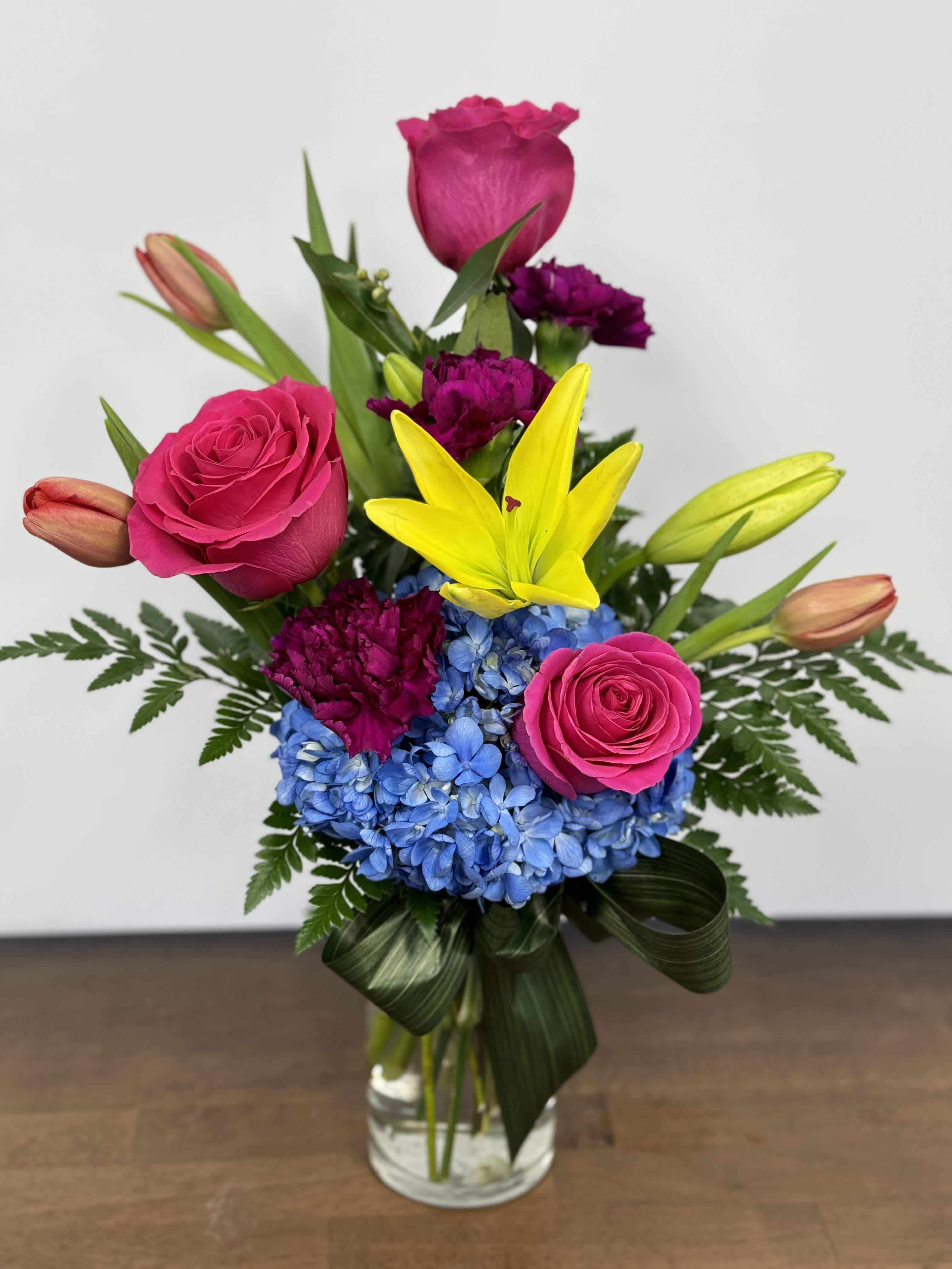 Bright bouquet with pink roses, yellow lilies, blue hydrangea, and purple carnations in a glass vase