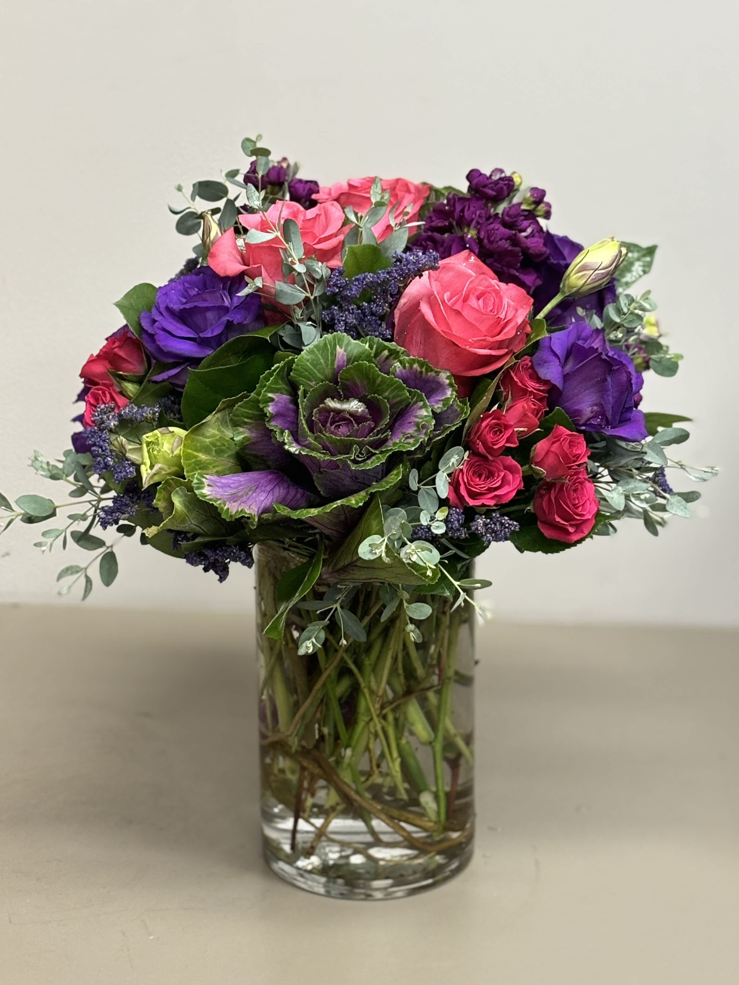 Vibrant arrangement of pink roses, purple flowers, and ornamental cabbage in a clear glass vase