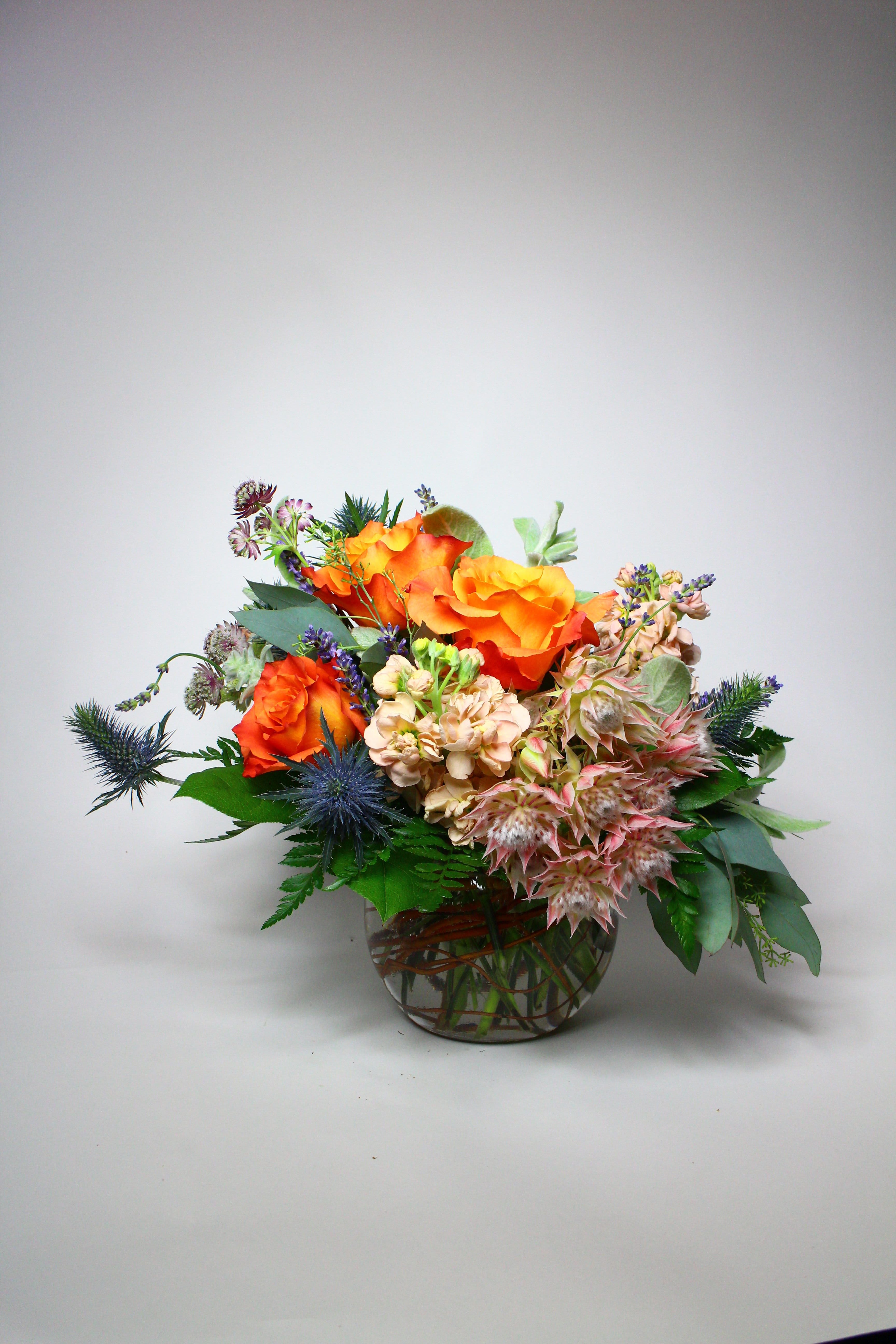 Orange roses arranged in a glass bowl with blue thistle and pale pink blooms