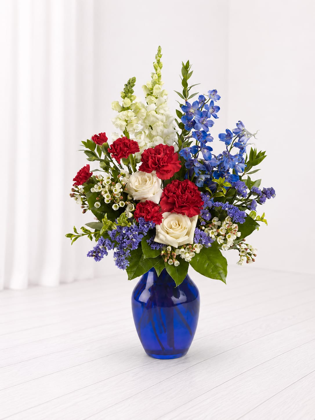Patriotic Peace - Honor a loved one with the timeless beauty of the Patriotic Peace Sympathy Bouquet. Designed in a striking cobalt blue vase, this arrangement features rich red carnations, crisp white roses and snapdragons, and vibrant blue delphinium accented with soft seasonal blooms and lush greenery. With its classic red, white, and blue palette, this thoughtful tribute offers comfort, remembrance, and a meaningful expression of sympathy and honor.