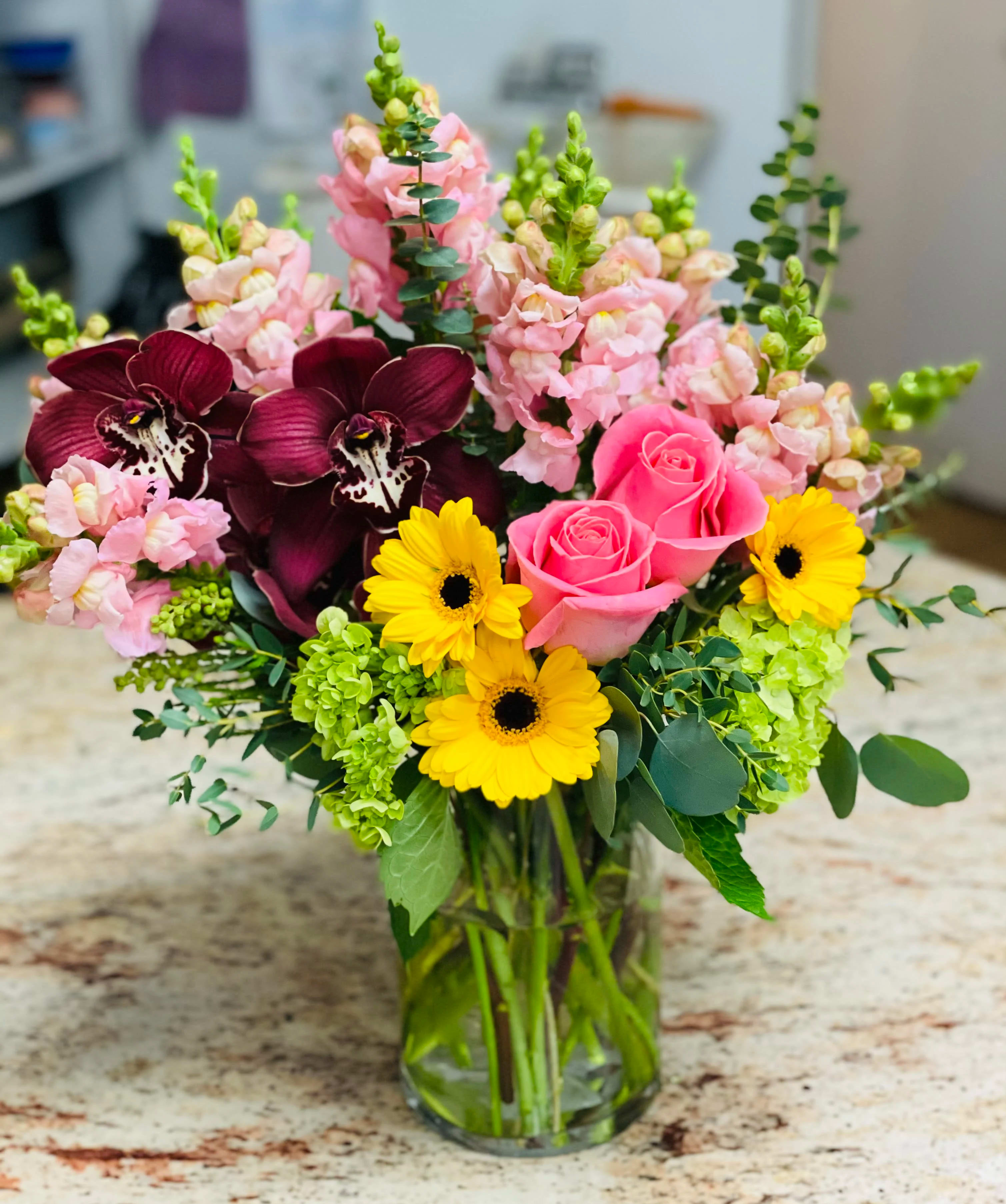 Mixed bouquet with pink roses, yellow daisies, and dark orchids in a glass vase