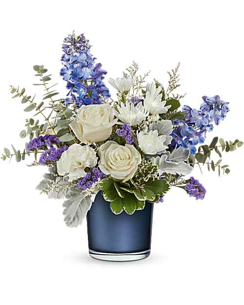 Sapphire Bliss - Tranquil blues! This lovely crème rose bouquet resembles fluffy white clouds against a clear blue sky, elegantly paired with a sapphire blue glass vase that shimmers with a pearlescent finish. This arrangement features crème roses, blue delphinium, white carnations, white cushion spray chrysanthemums, sinuata statice, and limonium, accented with dusty miller, spiral eucalyptus, and variegated pittosporum. 