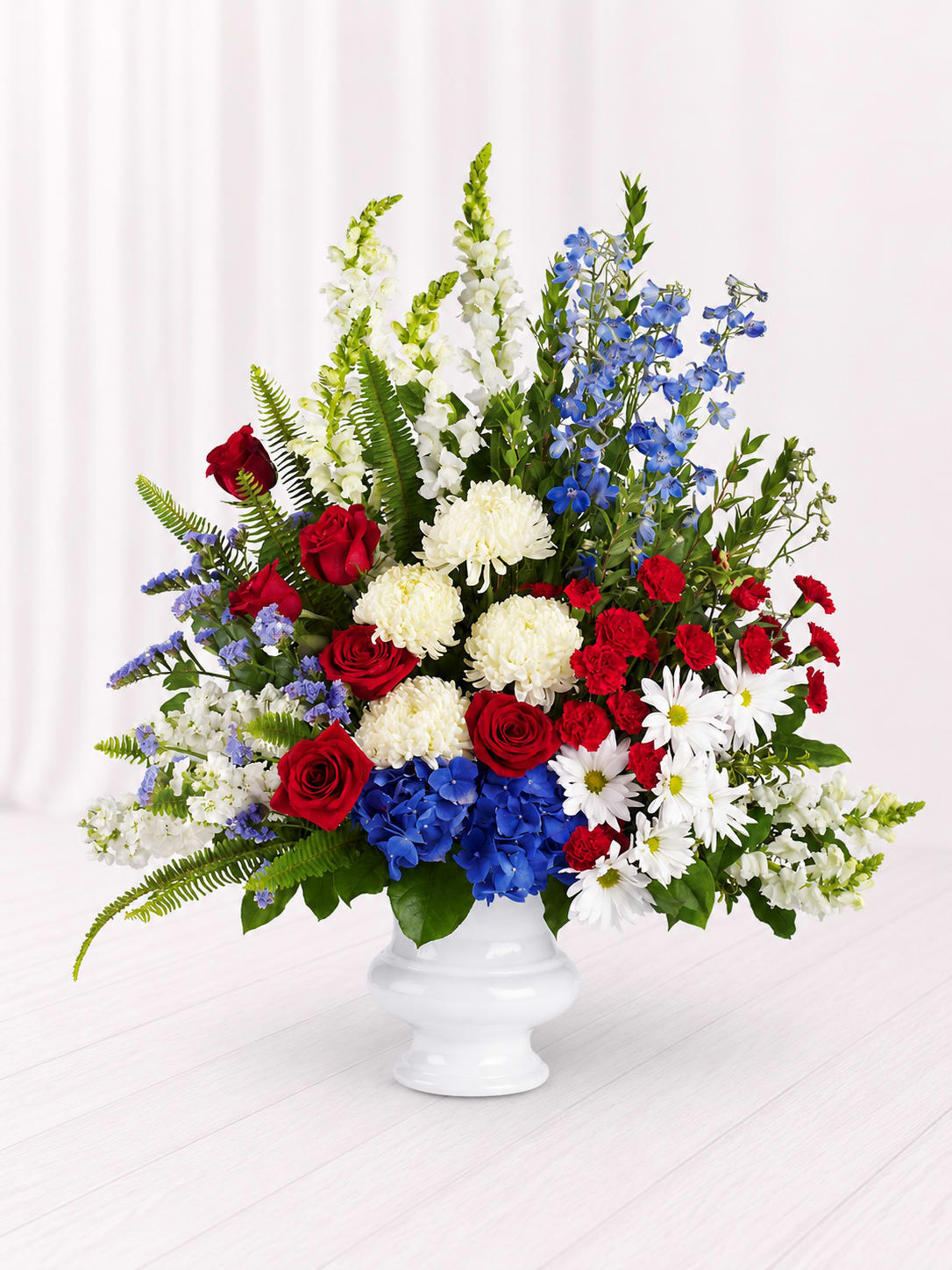 Honor &amp; Remembrance - A beautiful tribute of love, honor, and lasting remembrance, the Honor &amp; Remembrance Sympathy Arrangement features rich red roses, crisp white blooms, vibrant blue hydrangea and delphinium, and lush greenery artistically designed in a classic white urn vase. With its patriotic red, white, and blue palette, this elegant arrangement offers comfort and heartfelt support while honoring a life well lived.