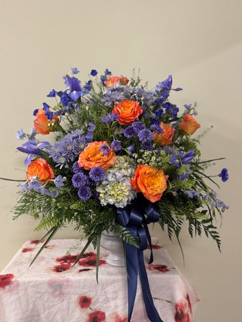 Celestial Comfort - Bright orange roses paired with vivid blue and purple blooms create an unforgettable pop of complementary color in this flower arrangement.  The arrangement blends roses, asters, hydrangeas and other seasonal filler flowers for an airy, textured silhouette with stems and greenery that extend outward for a natural, relaxed look. It is presented in a clean white urn vase finished with a long navy ribbon for a refined contrast that elevates the bright palette.  Note: Each item is subject to change in order to ensure the most beautiful and freshest blooms.