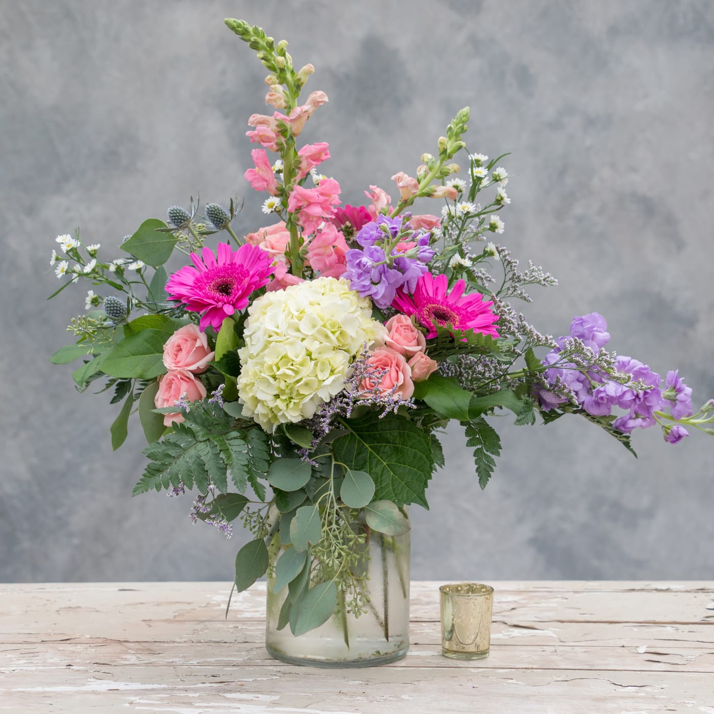 Mixed pink, purple, and white flowers in a glass vase beside a small candle holder.